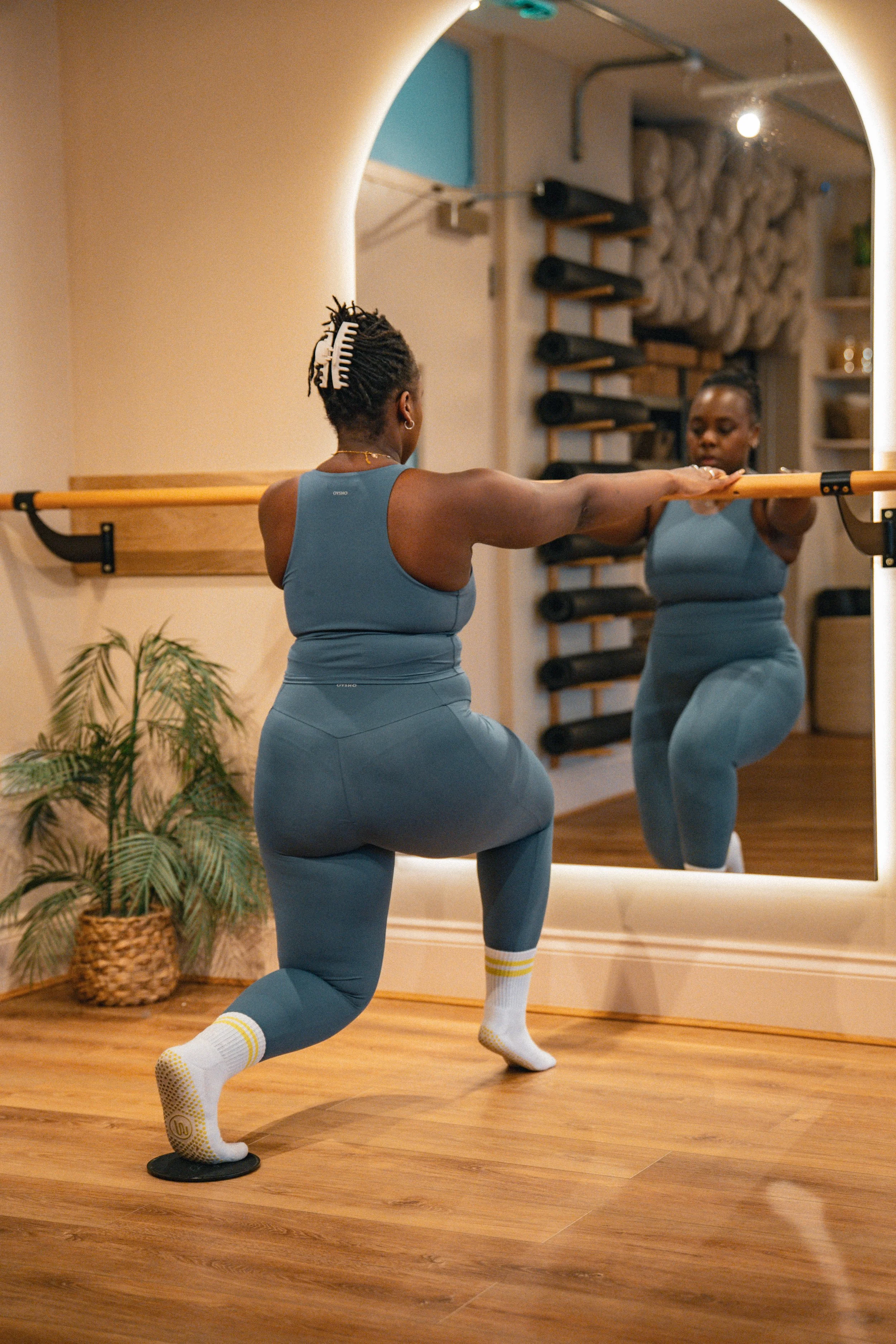 A person holding a ballet barre mounted on a wall with wooden paneling in a room with wooden floors and a potted plant in the background.