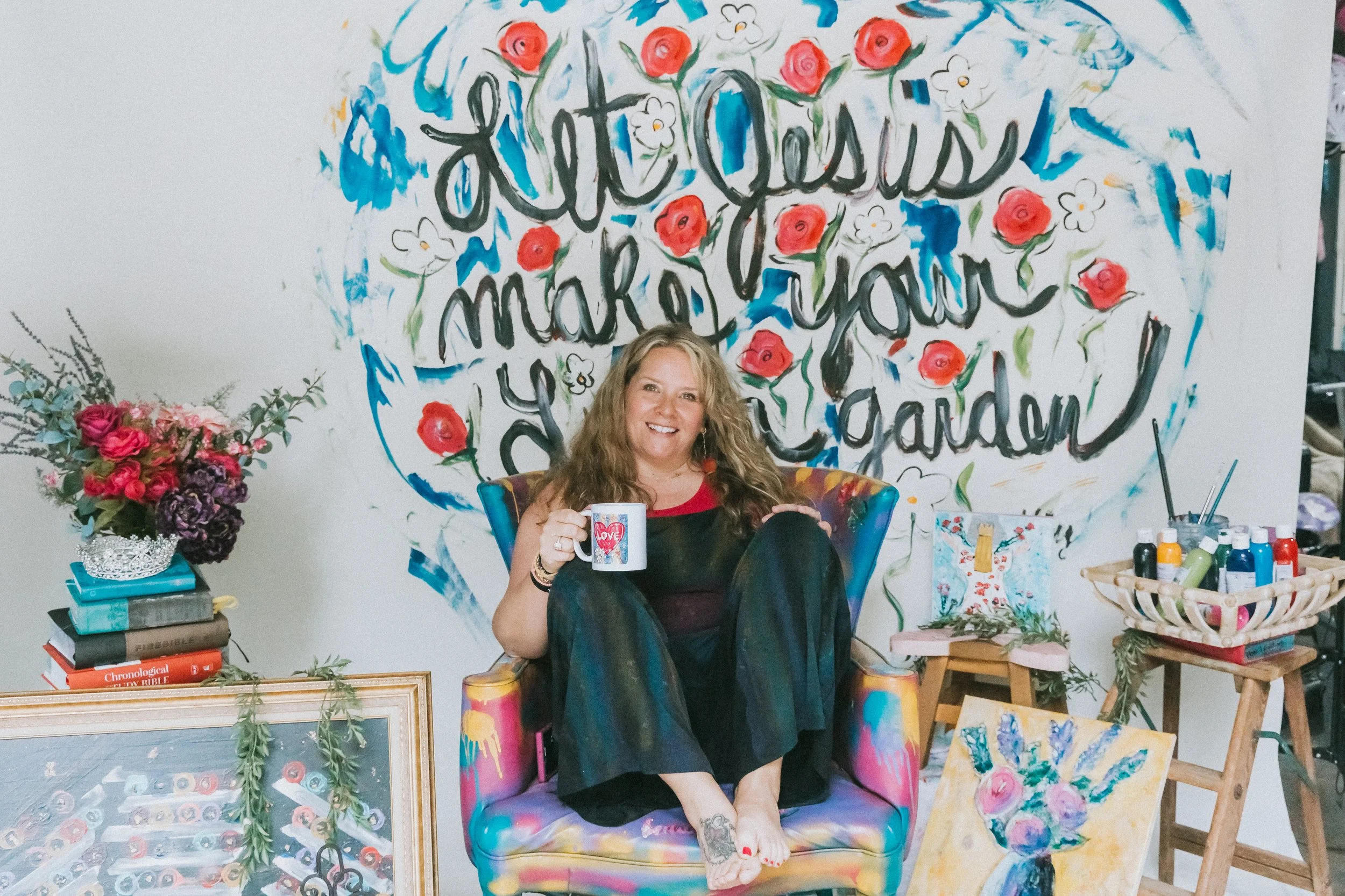 Jennifer Mehling sitting in a colorful painted chair with a large hand-painted mural behind her that reads “Let Jesus make your life a garden,” surrounded by art supplies and paintings.