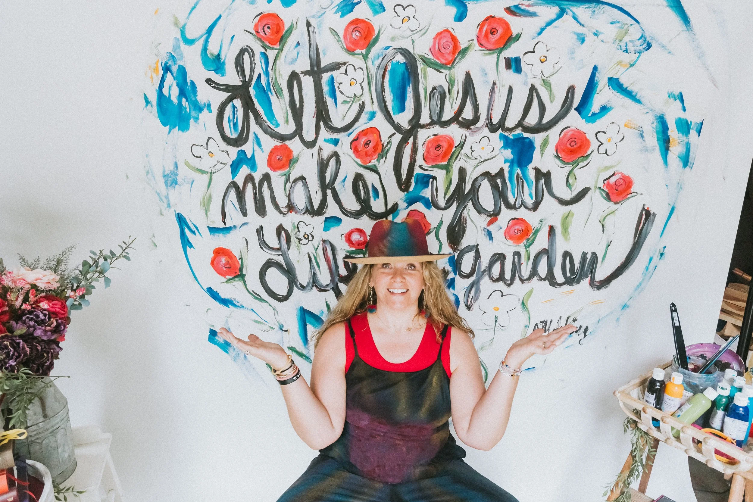Jennifer Mehling standing in front of a hand-painted wall that reads “Let Jesus make your life a garden,” gently covering her face with a colorful hat in a moment of surrender and reflection.