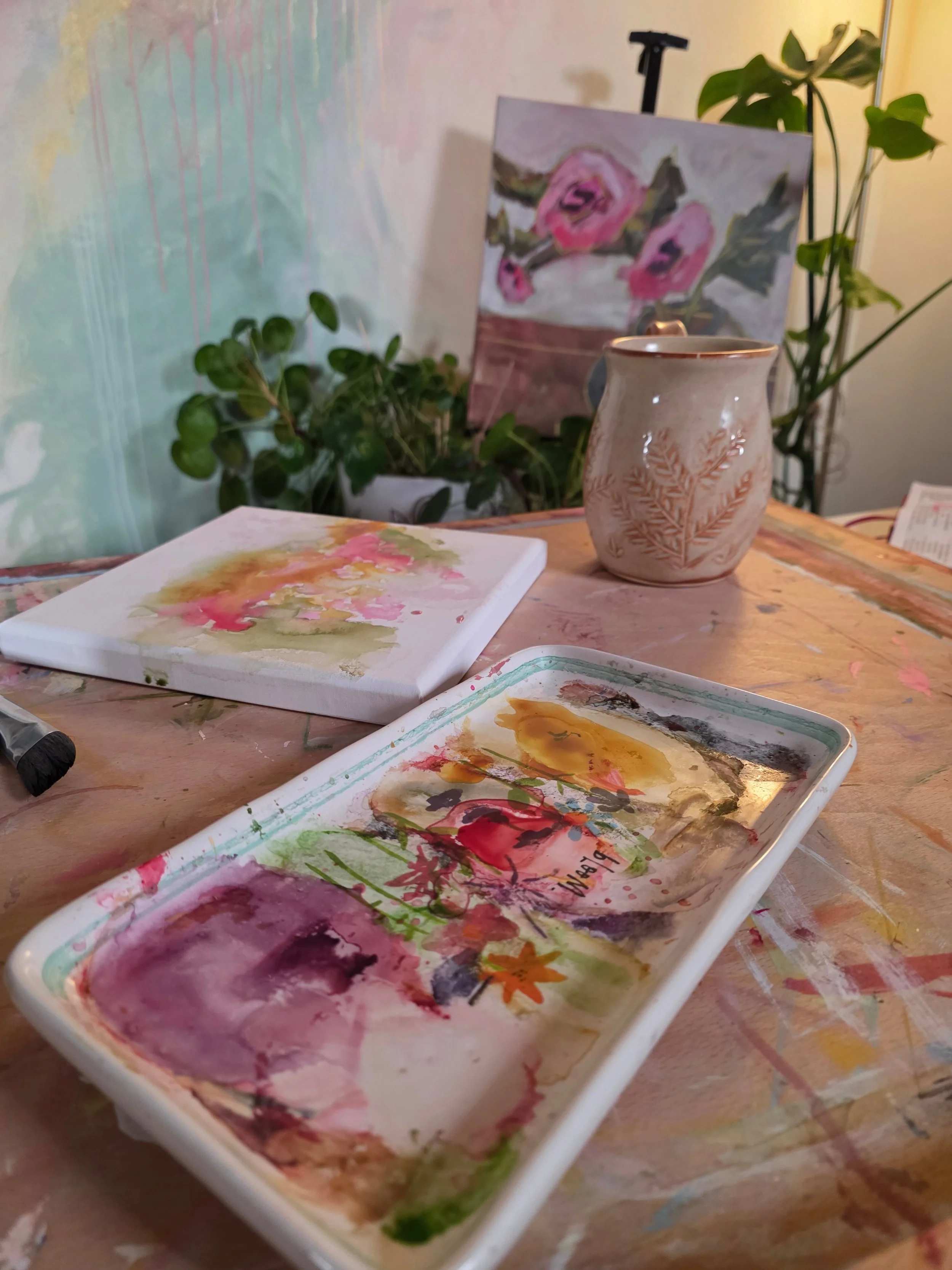 Watercolor palette, coffee mug, plants, and artwork on Jennifer Mehling’s studio table for the First Blooms collection.
