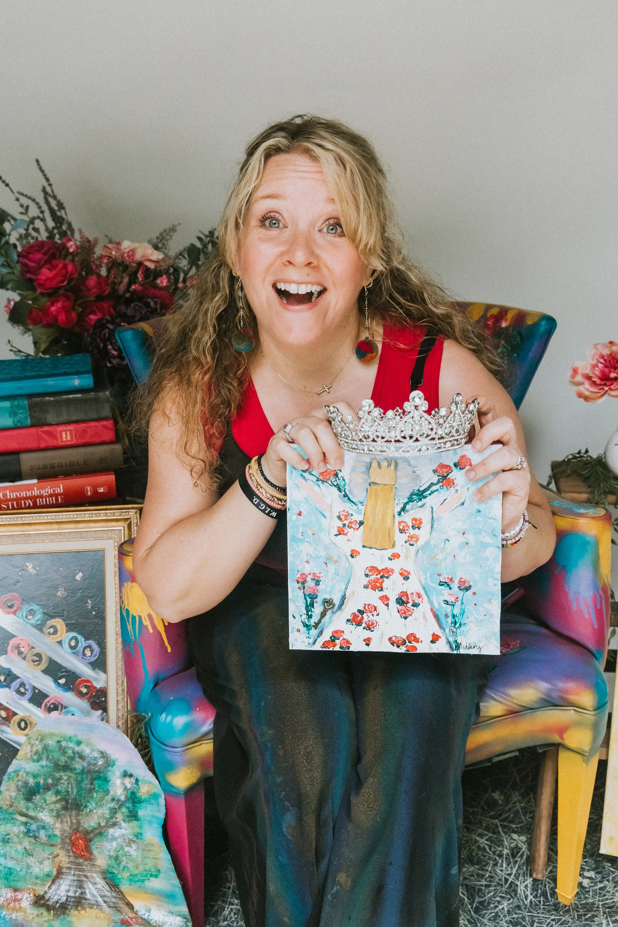 Jennifer Mehling smiling and holding a floral dress painting with roses, seated in a colorful chair surrounded by artistic decor.