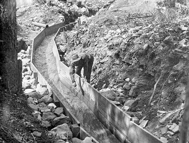 Black-and-white photograph of a miner in a hat working beside a long wooden sluice on a hillside during the California Gold Rush