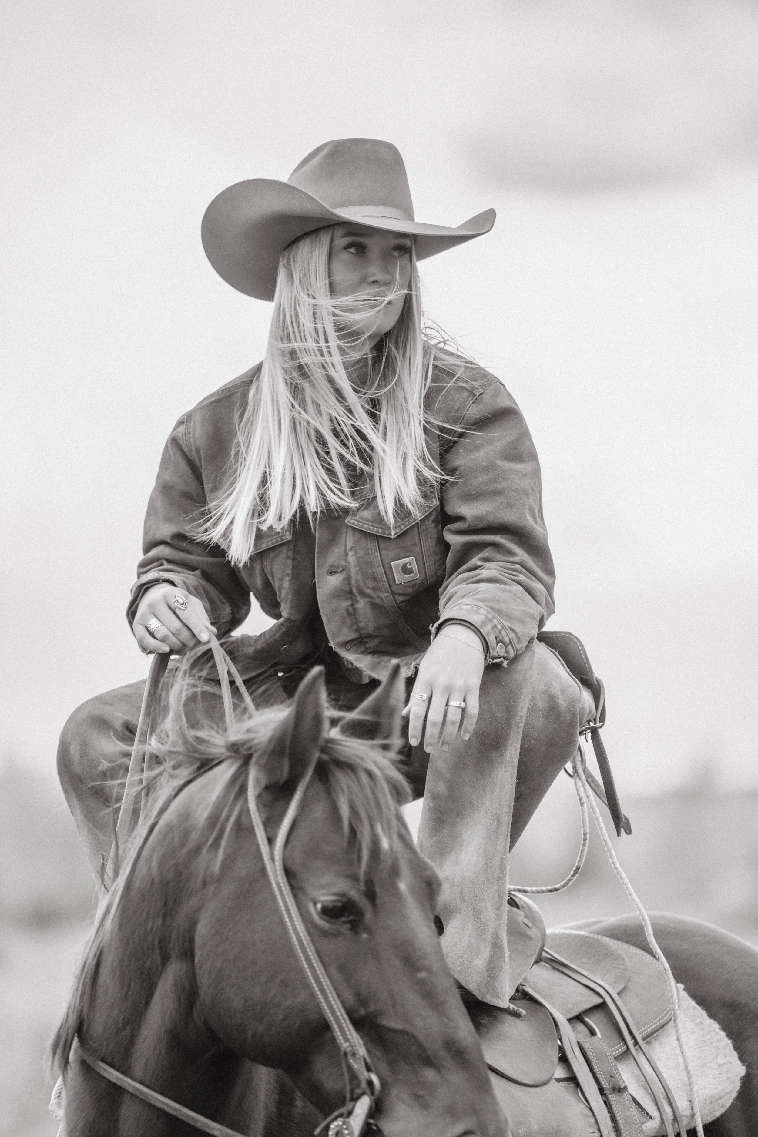 A woman wearing a cowboy hat and a denim jacket riding a horse in an outdoor setting.
