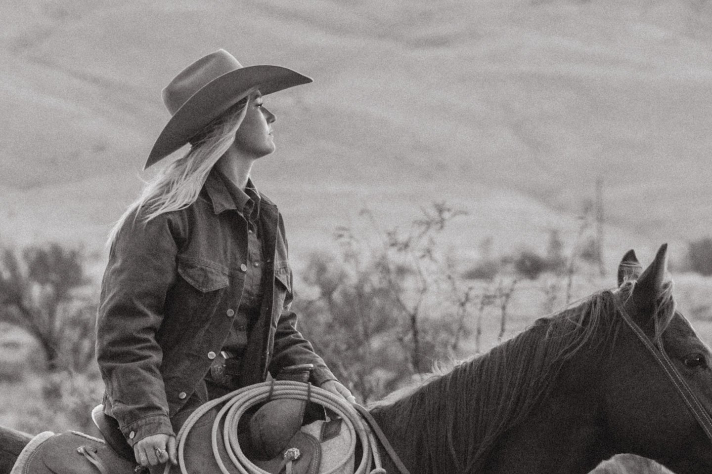 A woman riding a horse in a rural landscape, wearing a cowboy hat and denim jacket, black and white photo.