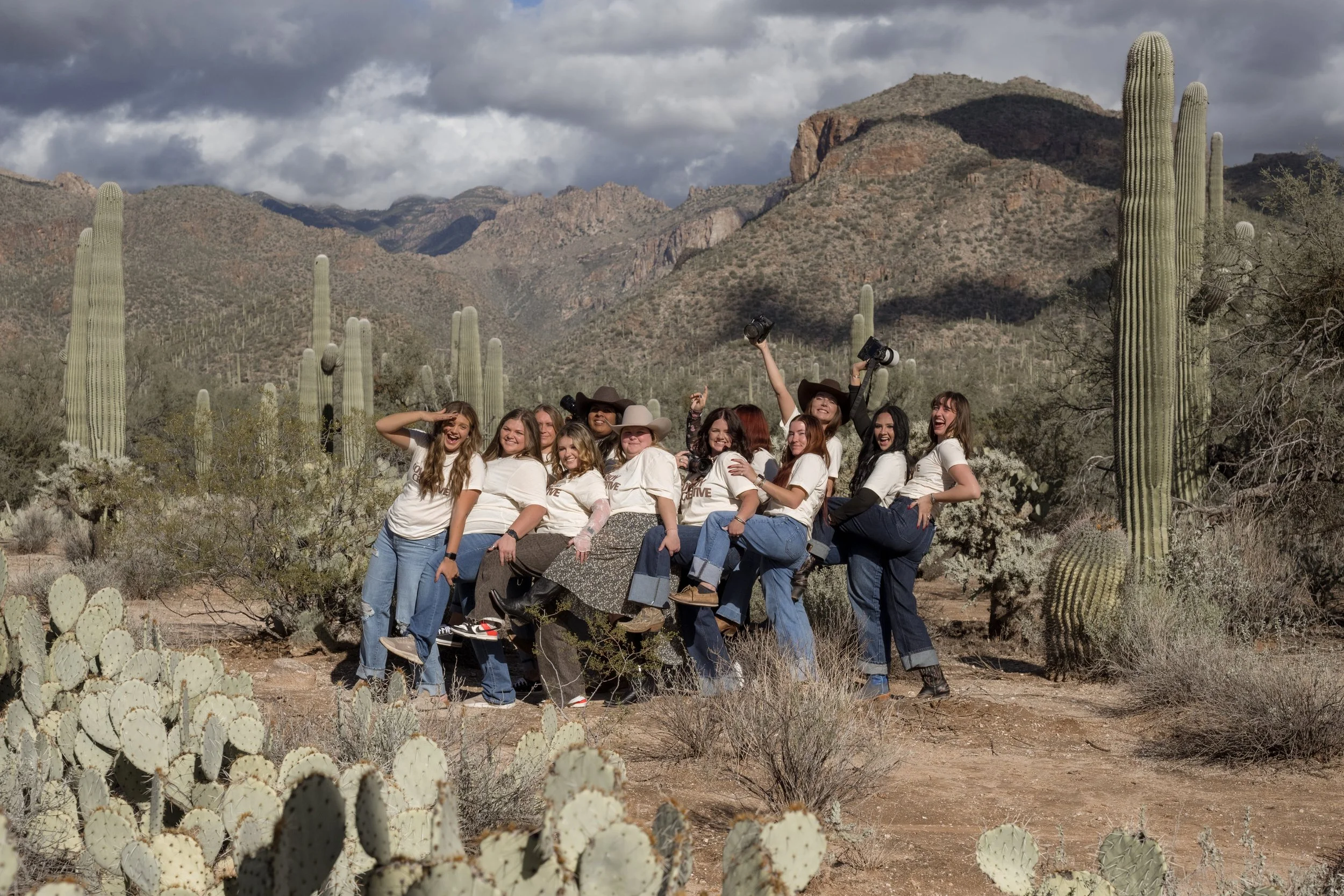 Group of women posing and smiling in a desert landscape with cacti and mountains, some wearing hats and holding cameras.