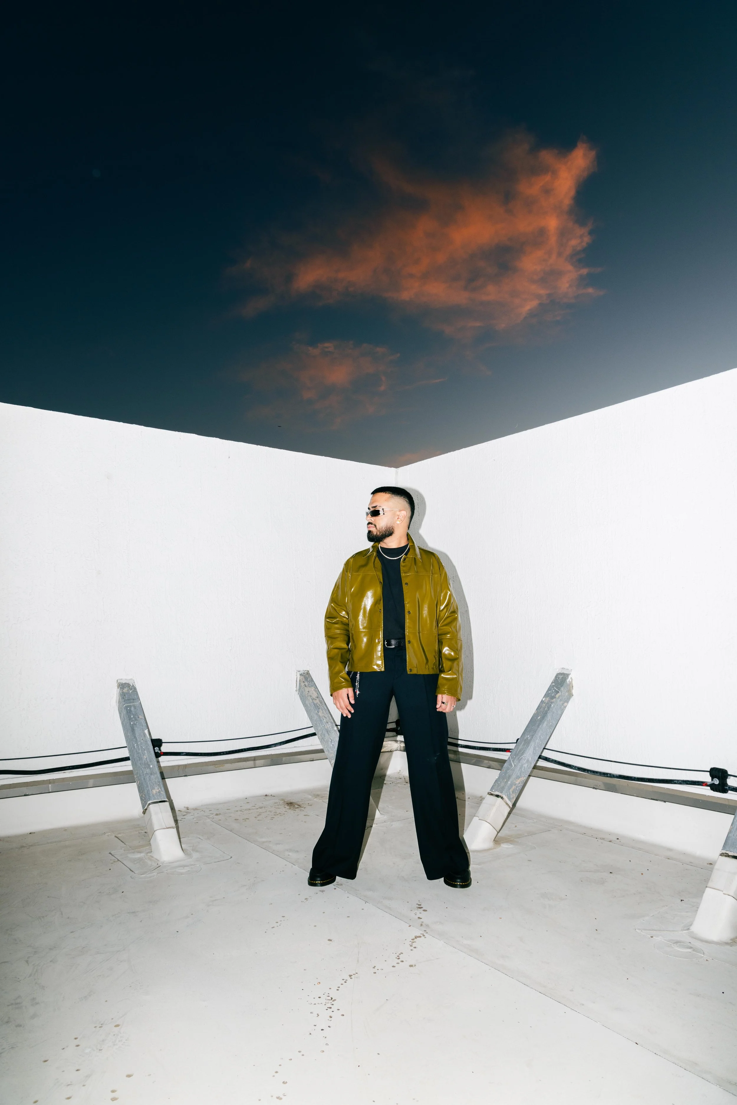 A man with sunglasses and a beard standing on a rooftop with a white wall, wearing a shiny olive-green jacket and black pants, under a dramatic sky with orange clouds.
