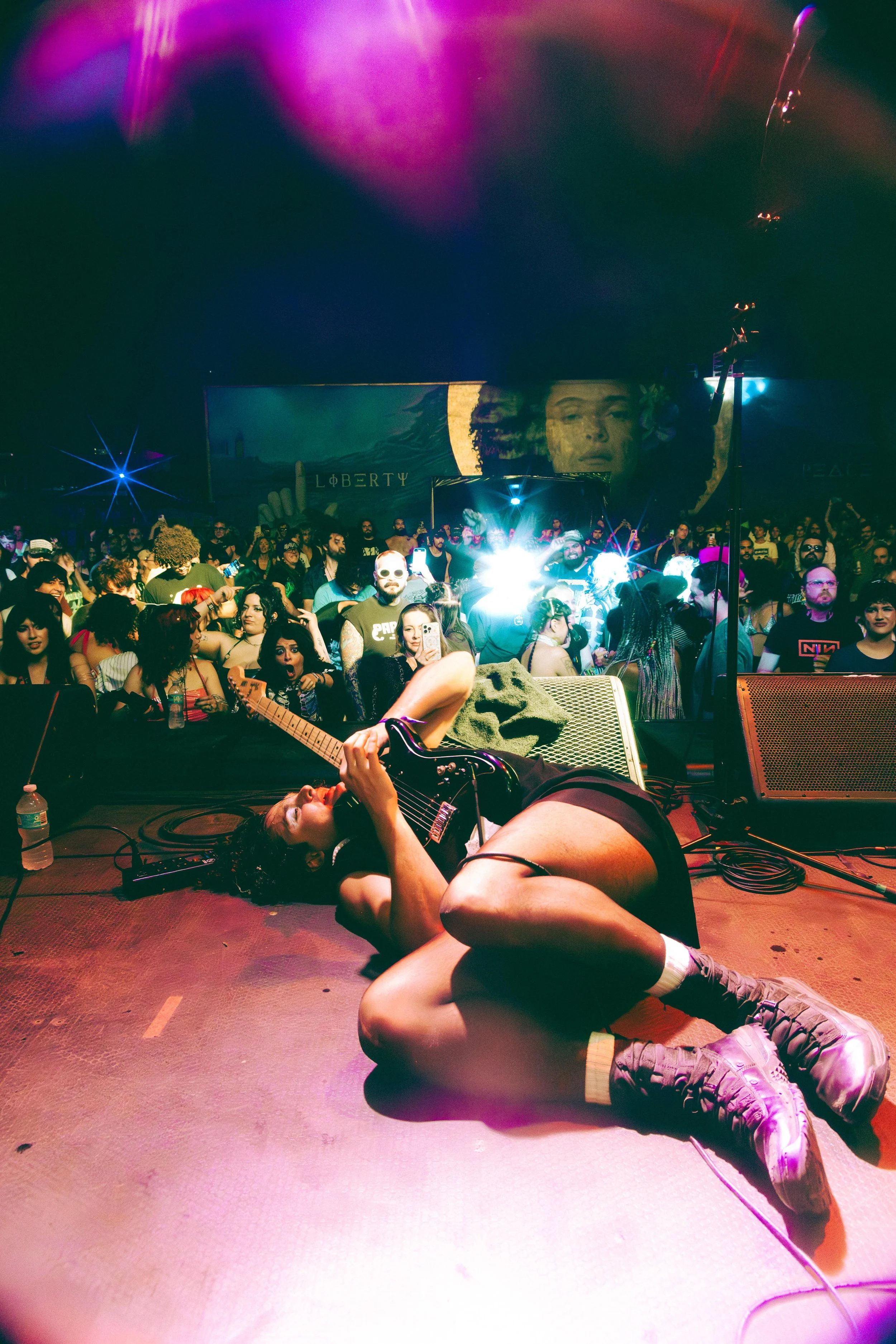 A female performer lying on the stage playing an electric guitar at a concert, with a crowded audience in the background and colorful stage lights overhead.