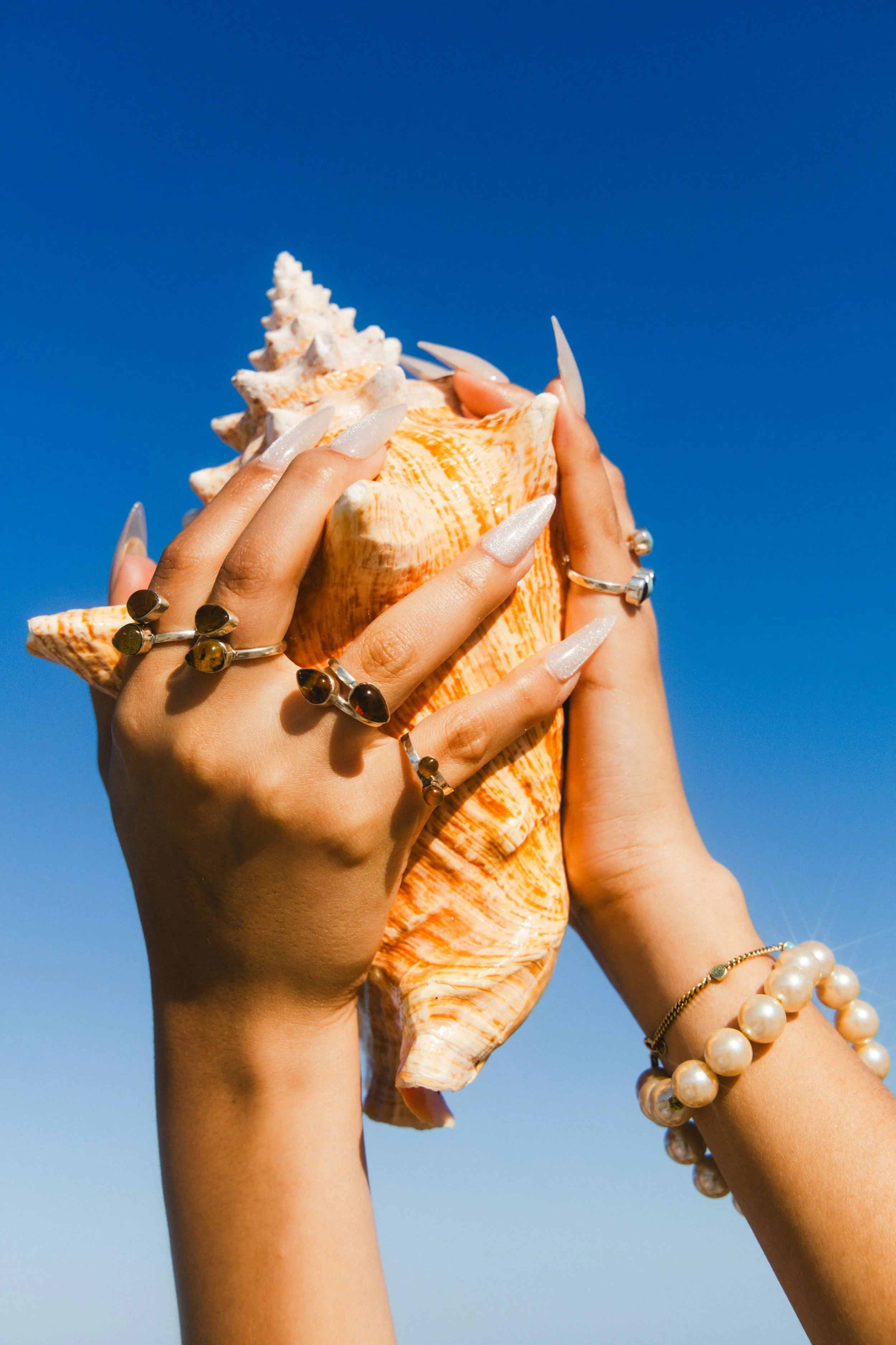 Hands holding a large seashell against a clear blue sky, adorned with rings, pearl bracelets, and long manicured nails.
