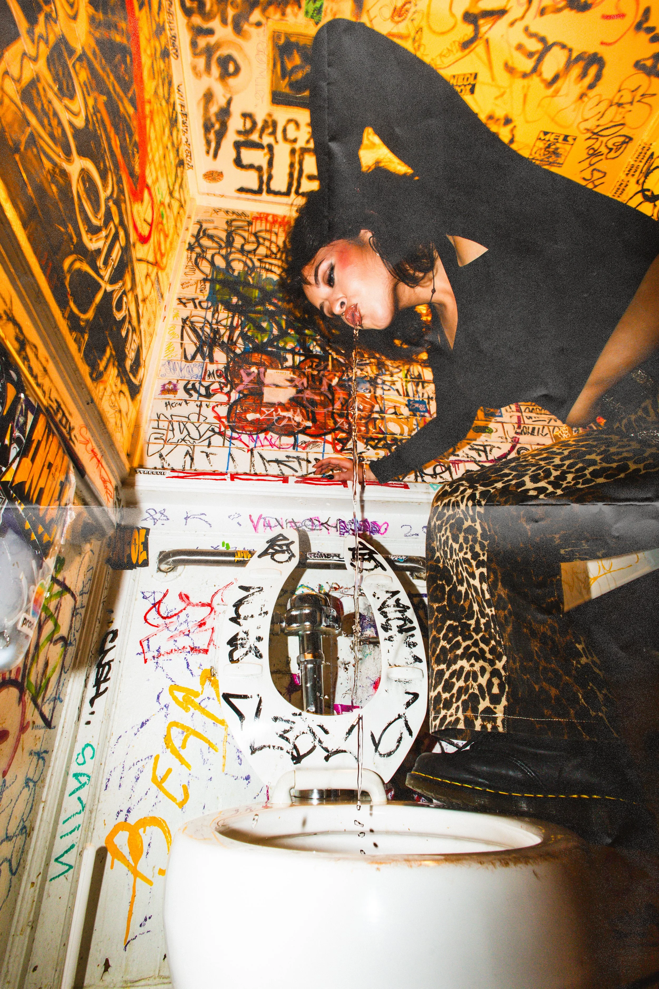 A woman with curly hair and makeup, wearing a black top and leopard-print pants, leans over a dirty bathroom toilet with graffiti-covered walls in the background, missing the toilet seat lid.