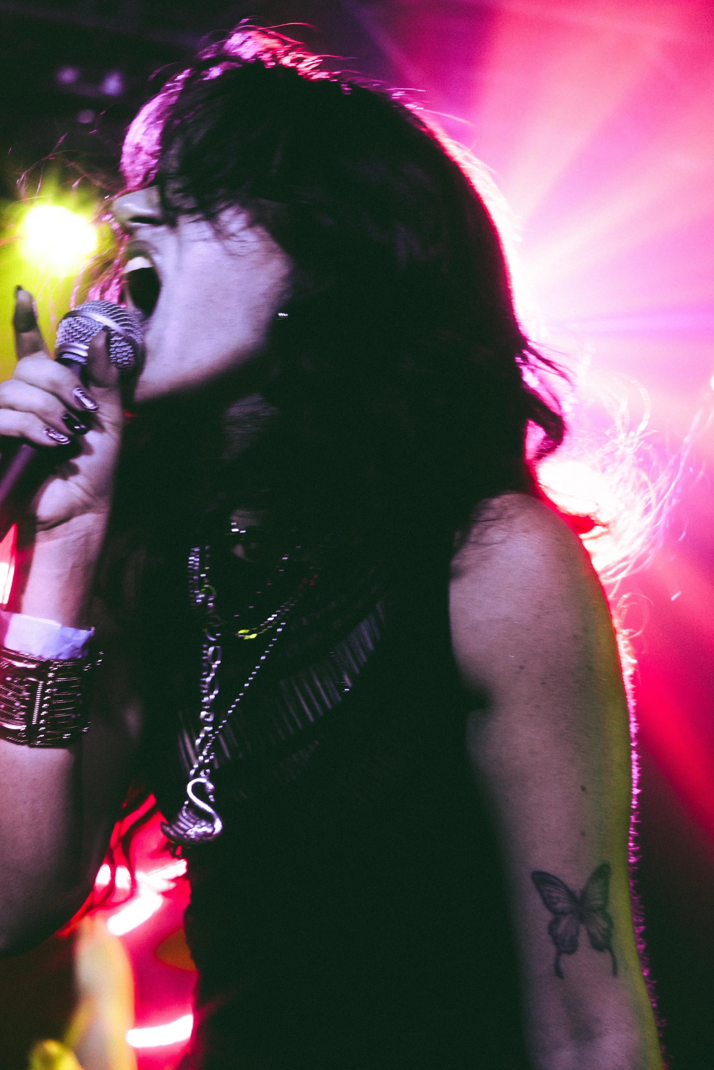 A woman with dark hair singing into a microphone on stage, with pink and purple stage lights illuminating her and a butterfly tattoo on her arm.
