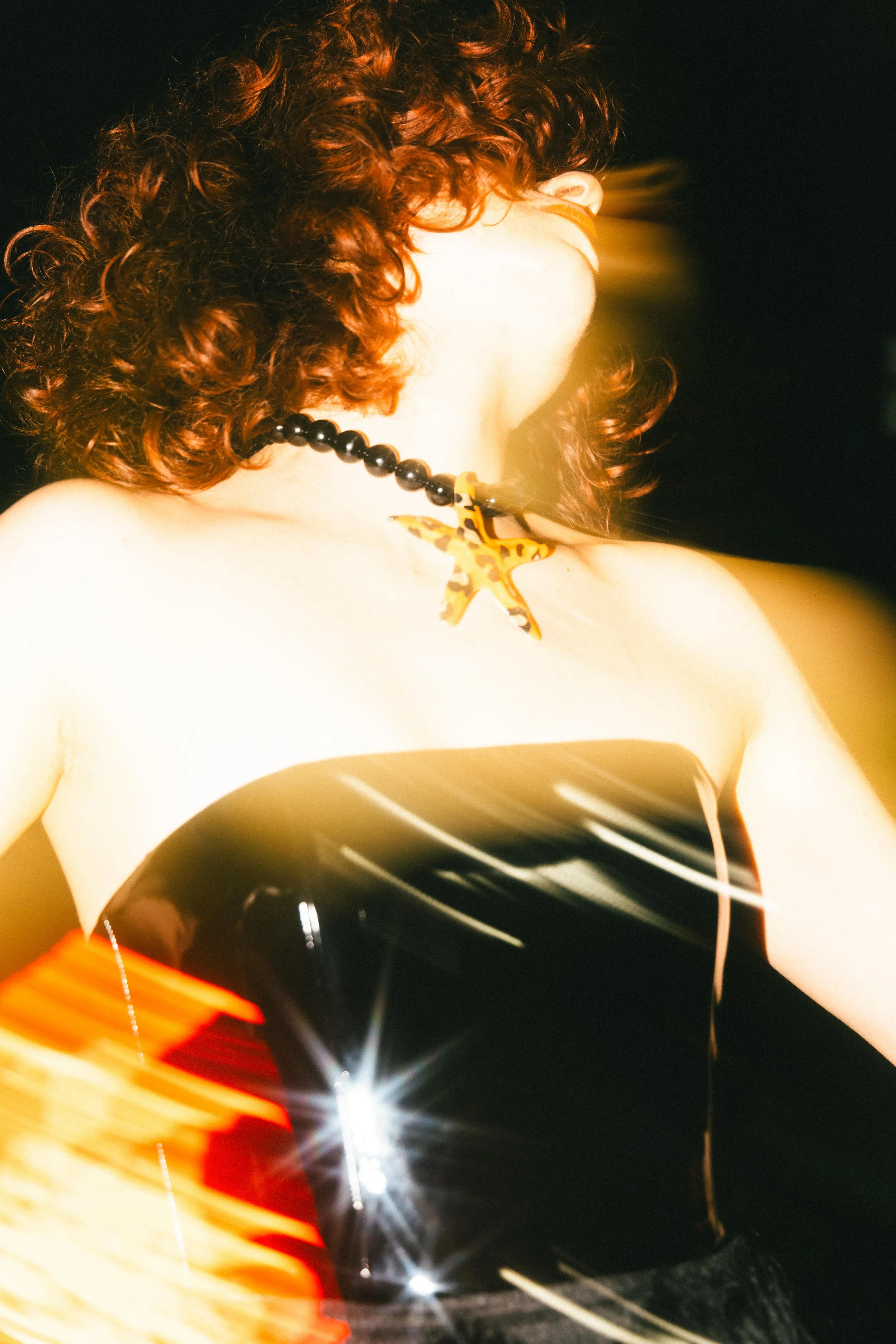 A person with curly red hair wearing a black strapless top, a beaded necklace with a butterfly pendant, and a bracelet with a yellow and black butterfly design, illuminated by bright, star-like light reflections.