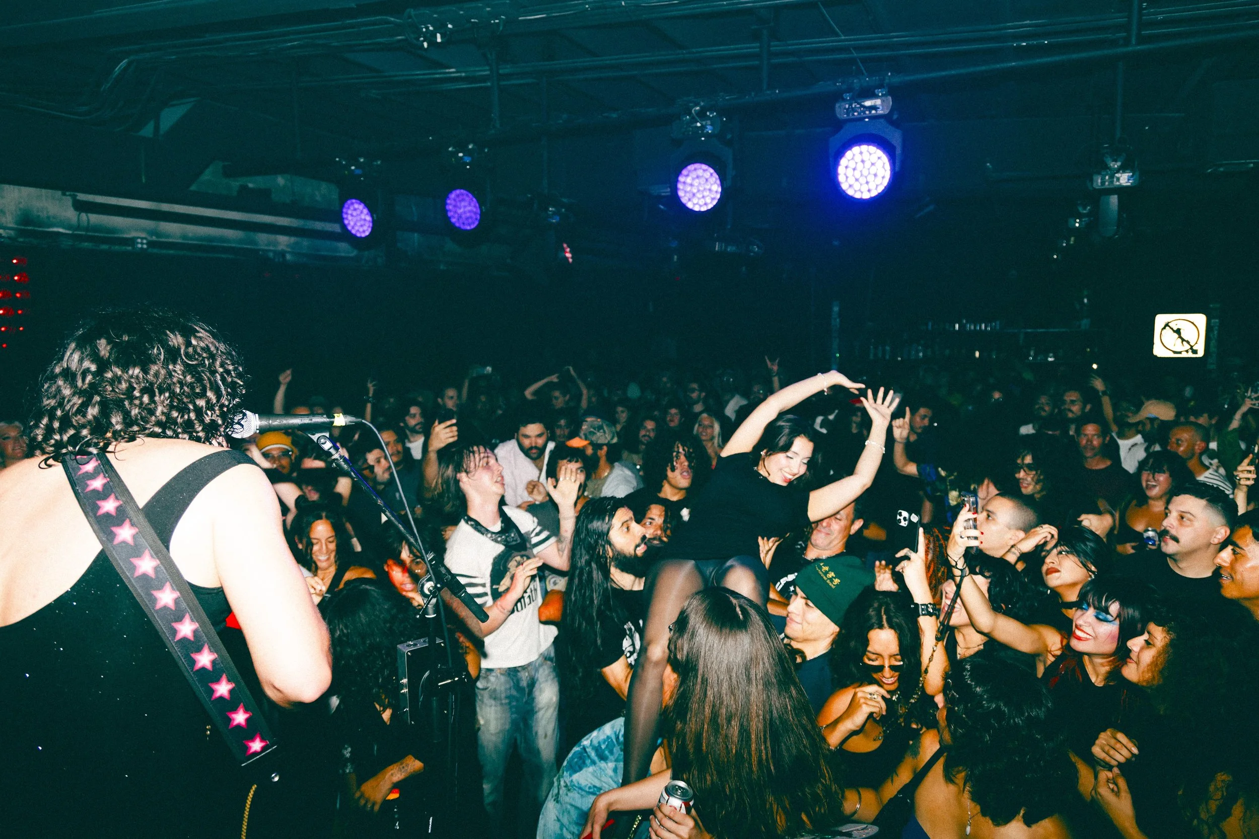 Crowd of people dancing and enjoying music at a concert or club with purple stage lights overhead.