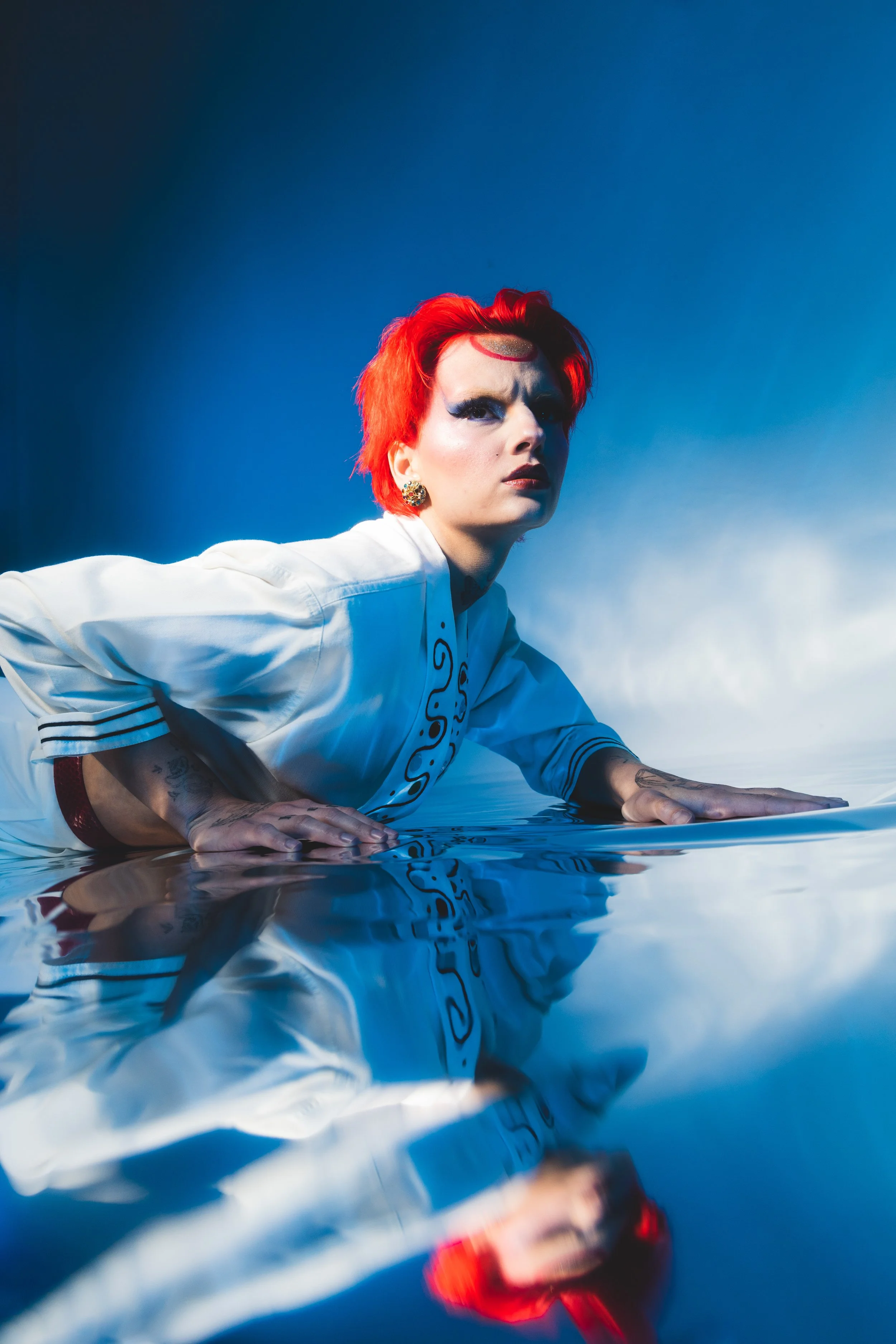 A person with bright red hair and bold makeup lying on a reflective surface, wearing a white top with black accents, against a blue background.