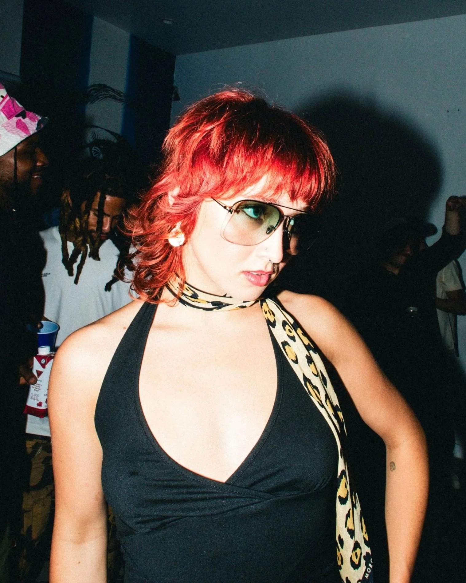 A woman with short red hair, wearing aviator sunglasses, a black top, a leopard print scarf, and large earrings, at a party or social gathering.
