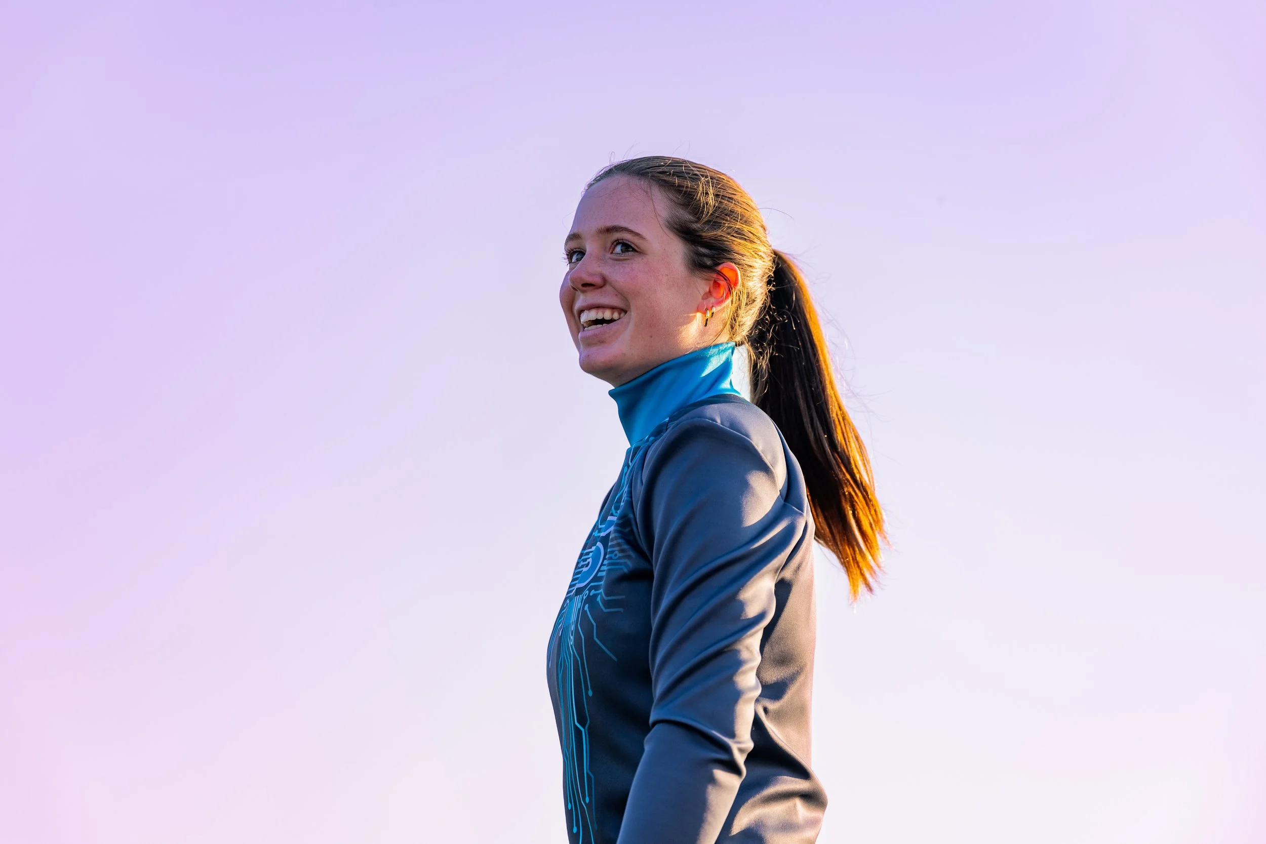 A young woman with long brown hair tied back, smiling and looking to the side, wearing a gray athletic jacket with blue accents and a blue turtleneck underneath, standing outdoors against a clear sky.