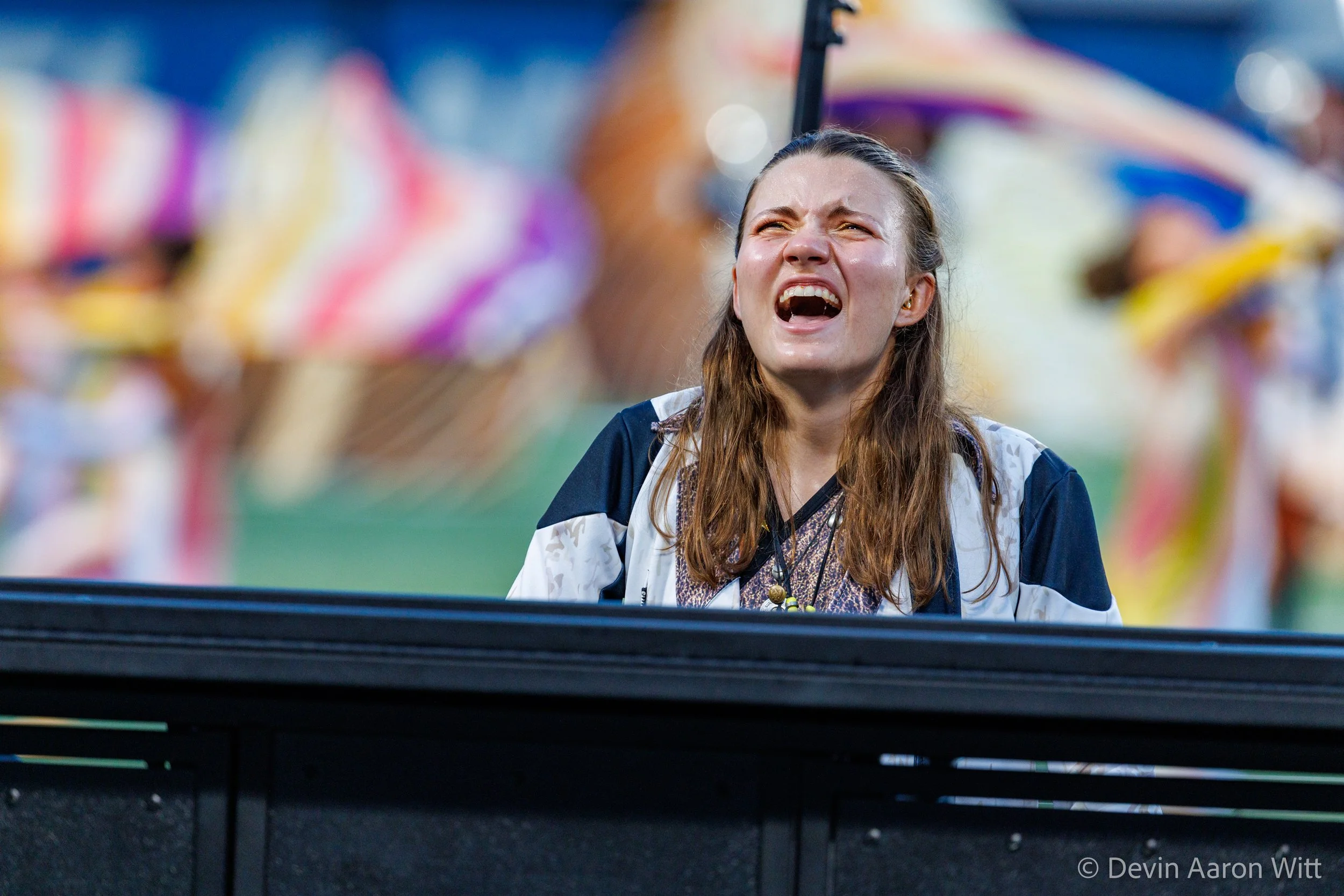 A young woman with long brown hair and a distressed expression, looking upward, standing behind a black barrier at a colorful fair or carnival with vibrant tents in the background.