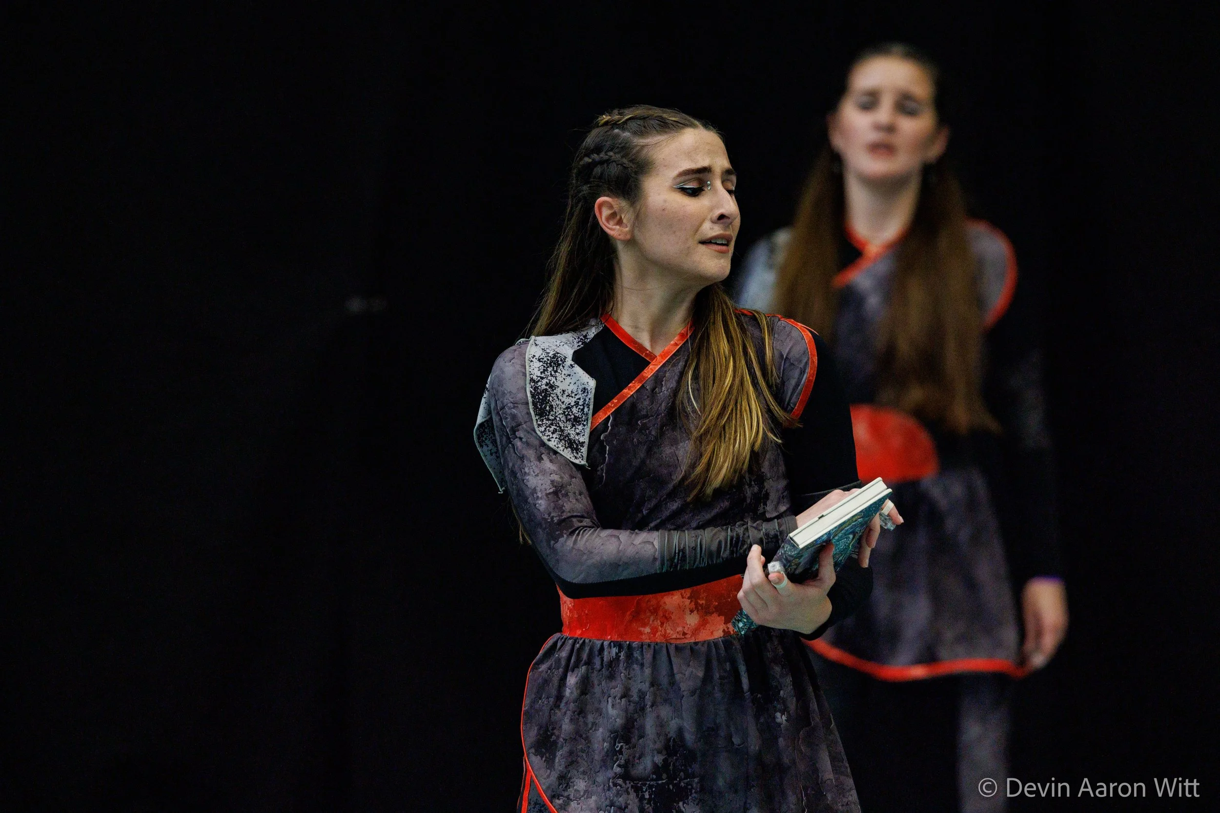 Two women on stage, one in the foreground holding a book, wearing a dark patterned dress with red accents, with an emotional expression, and the other in the background wearing a similar dress, slightly out of focus, against a black backdrop.