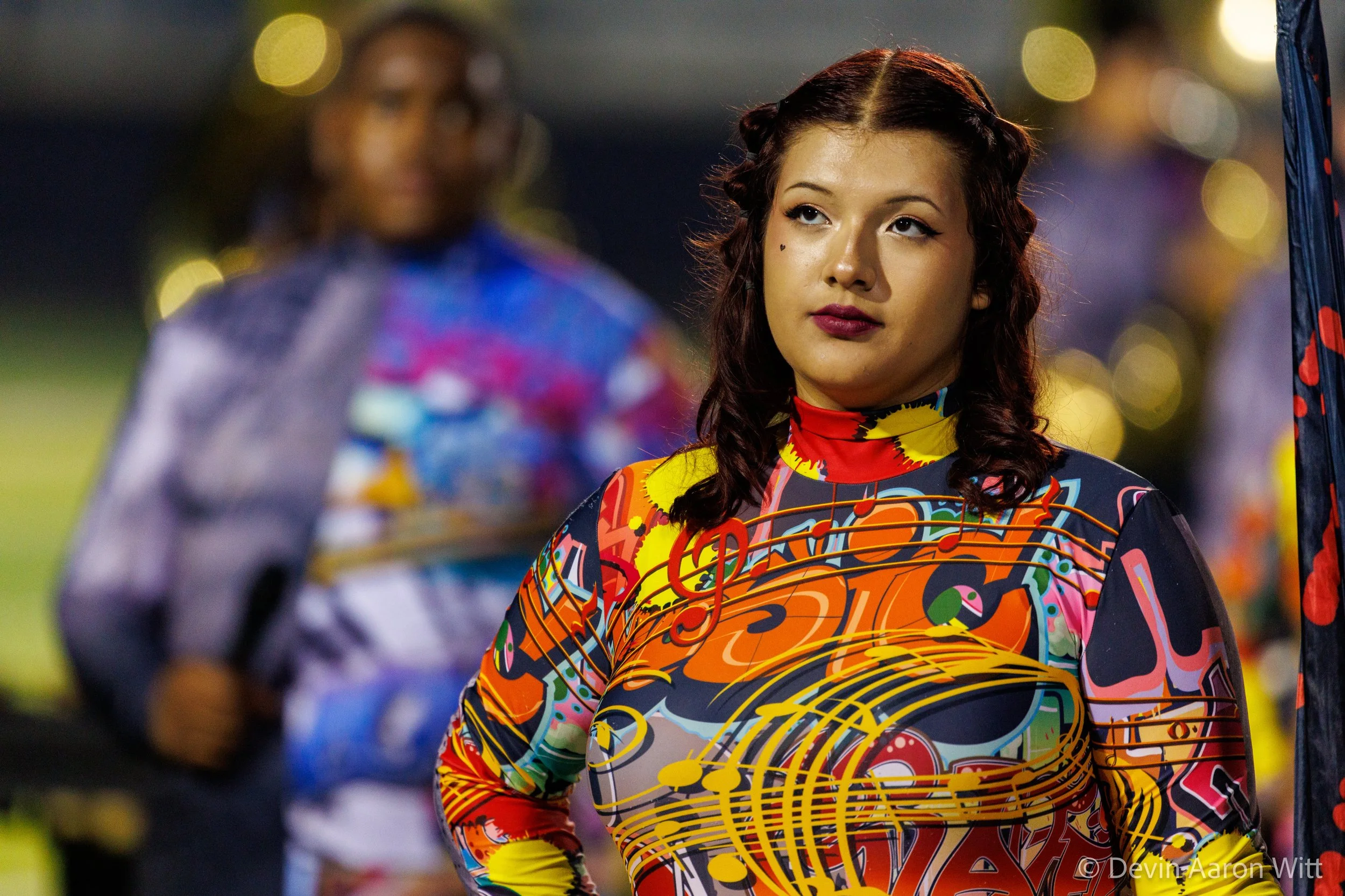 A woman with styled hair, wearing a colorful, musical note and swirl patterned long sleeve shirt, stands outdoors at night with blurred lights and a person in the background.