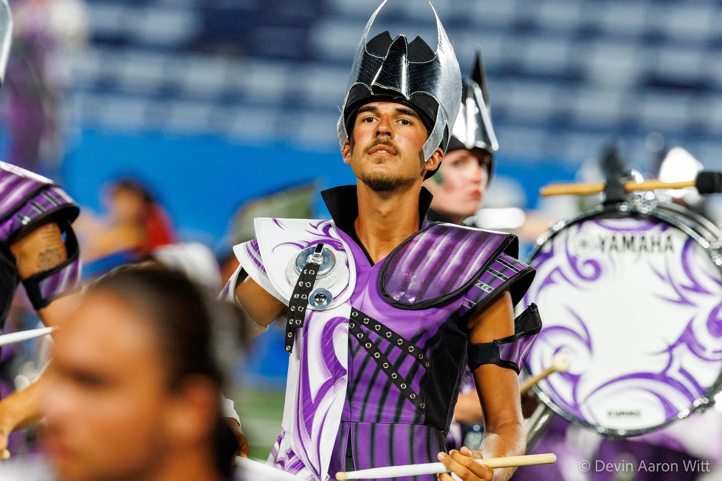 A marching band member in a purple and white uniform with a black and silver helmet holding a drumstick, with other band members and a large drum in the background.