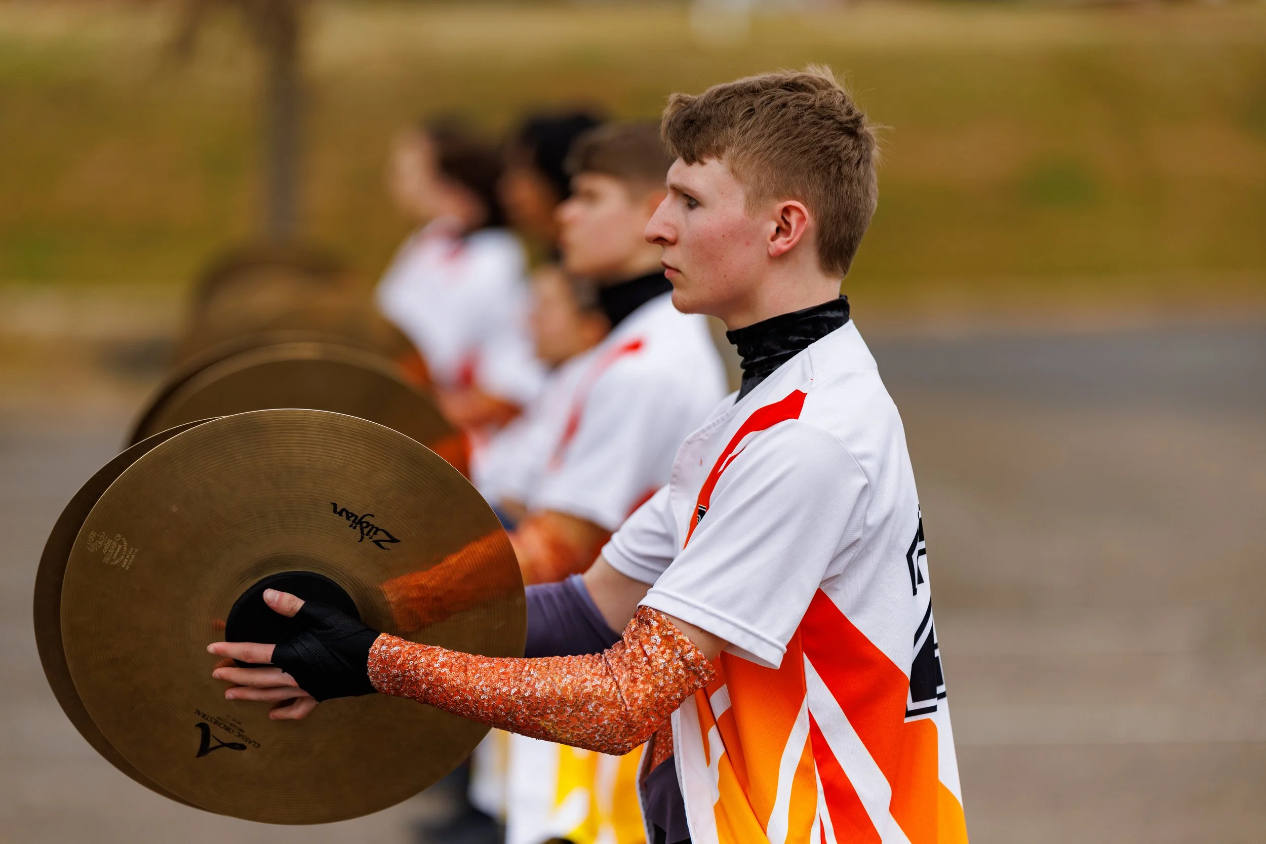 Young man in sports uniform holding a cymbal during an outdoor practice or event with teammates in the background.