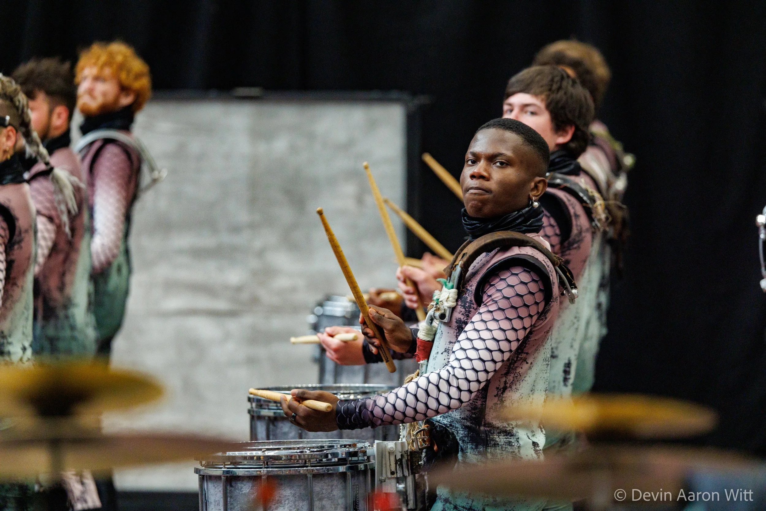 A young man playing drums with others in a band or orchestra, wearing a fishnet-style shirt and black scarf around his neck, with a focused expression.