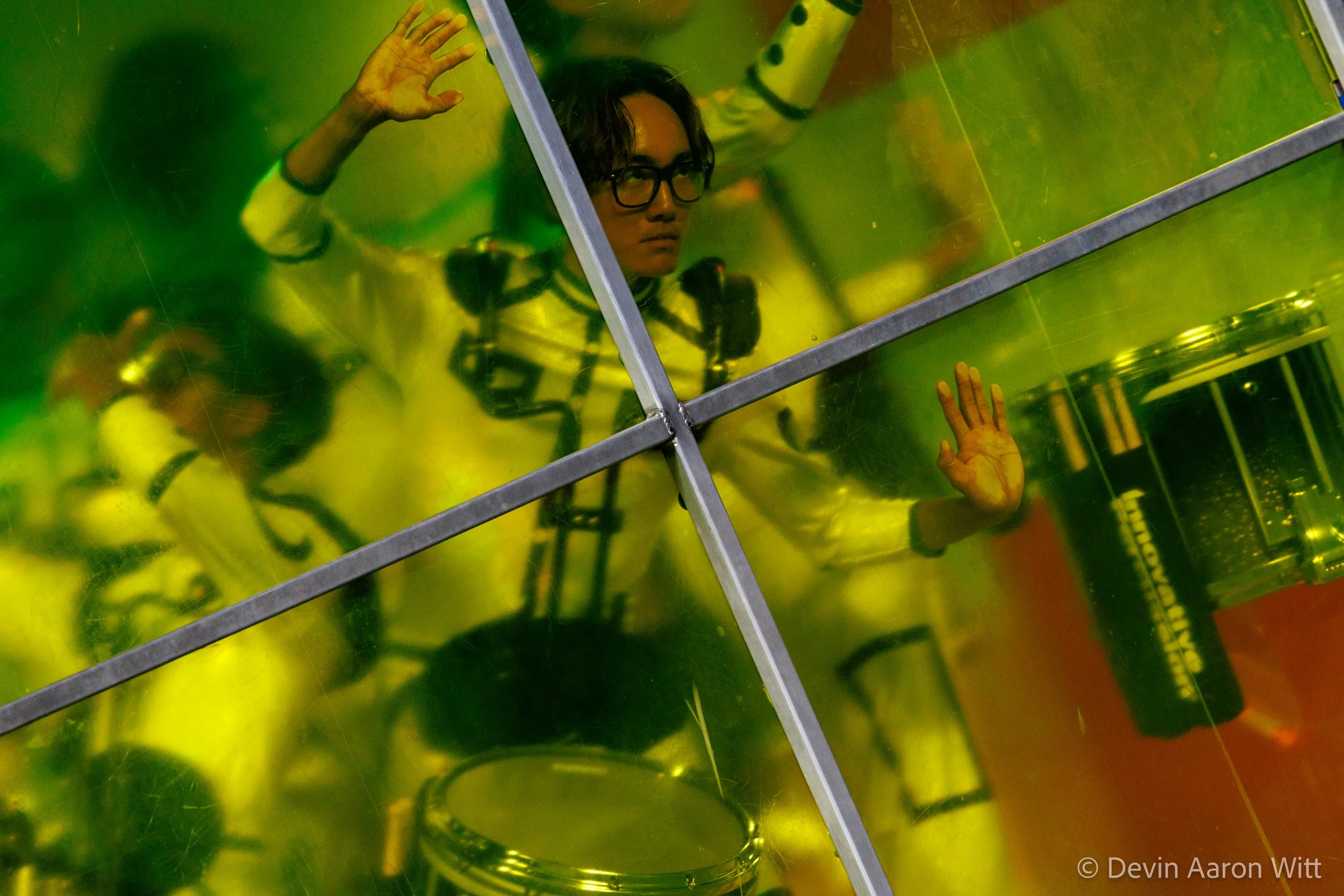 Person looking into a mirrored, yellow-tinted chamber, with hands pressed against the glass, surrounded by scientific equipment.
