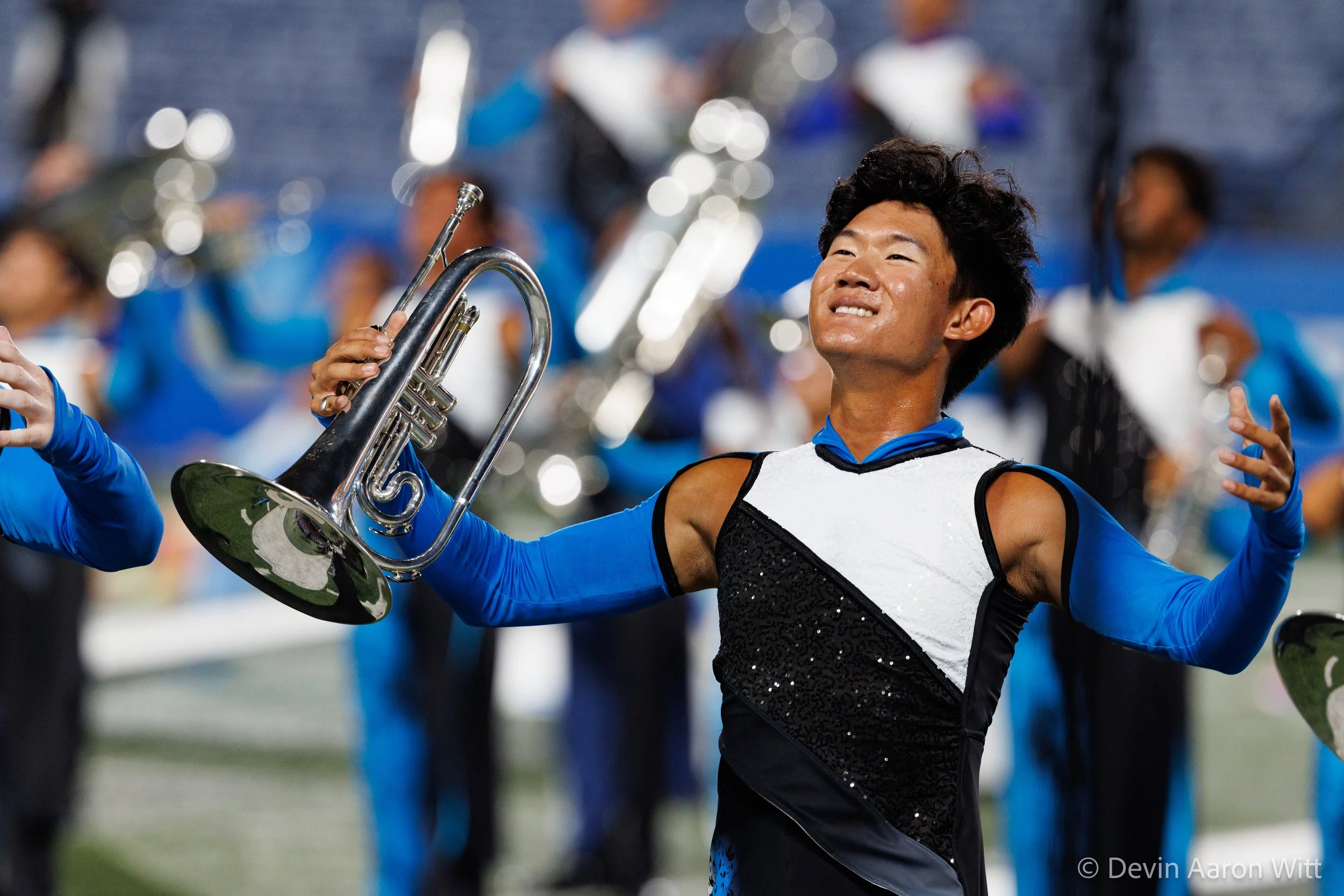 A young band member in a marching band holding a trumpet, smiling and appearing joyful during a performance, with other band members blurred in the background.