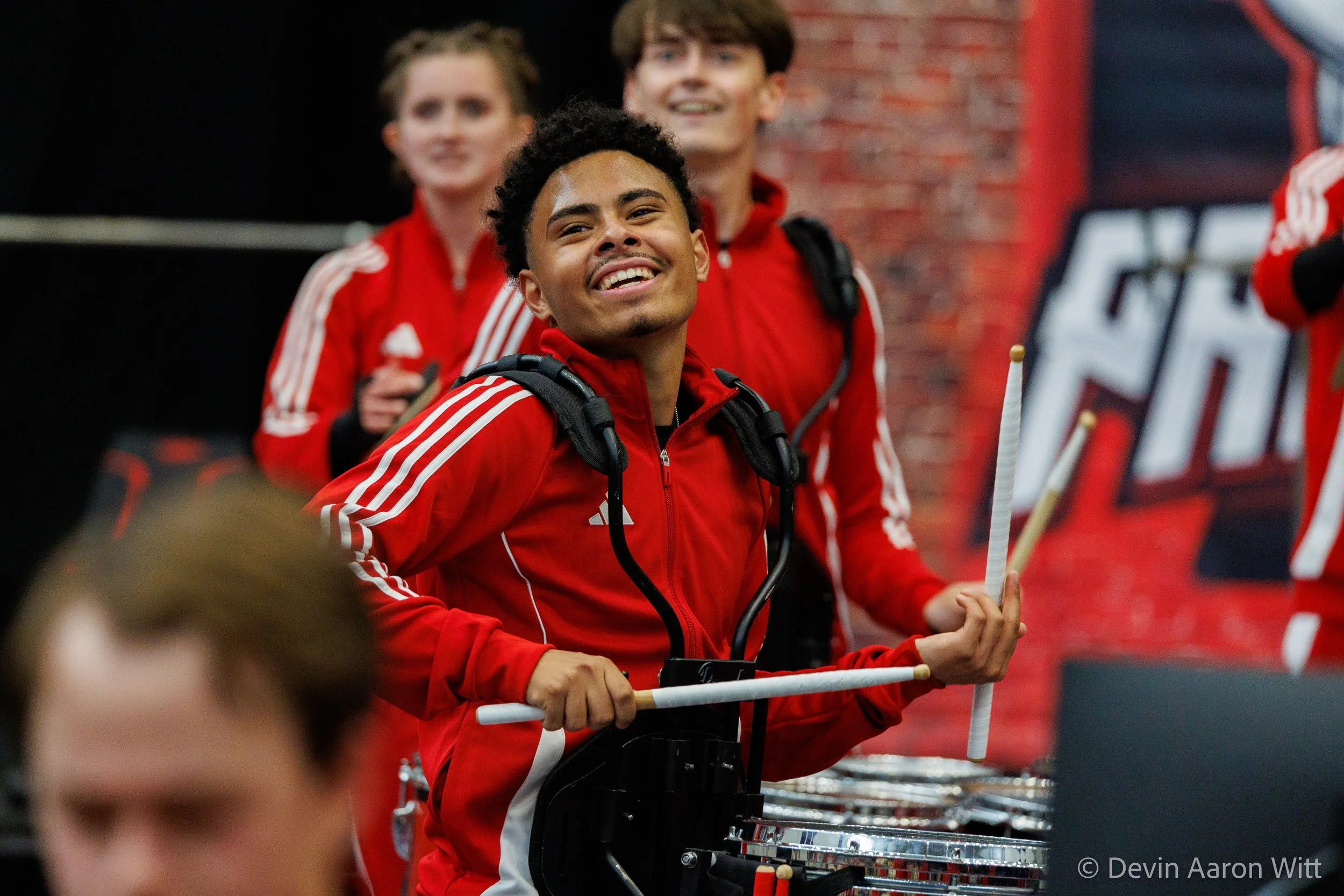 Young man in a red sports jacket smiling, holding drumsticks, with other young people in matching red sports jackets behind him, in an indoor setting with a brick wall and a banner.