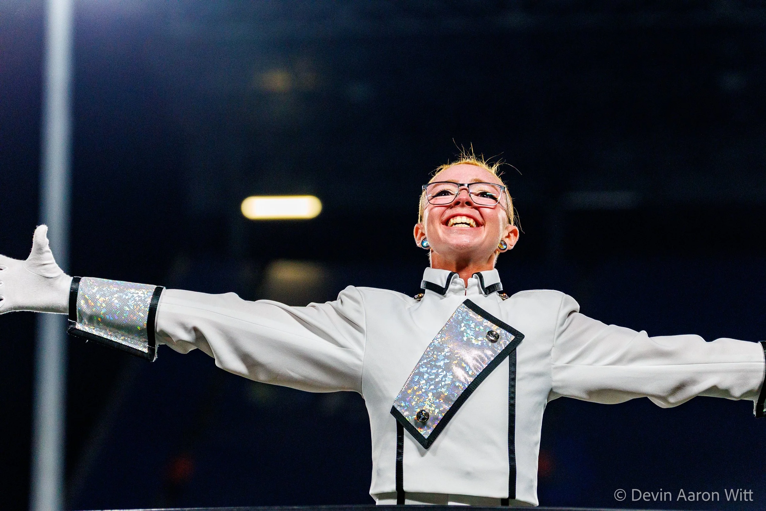A woman with glasses and earrings, wearing a white jacket with holographic accents, smiling with arms wide open in an indoor setting.