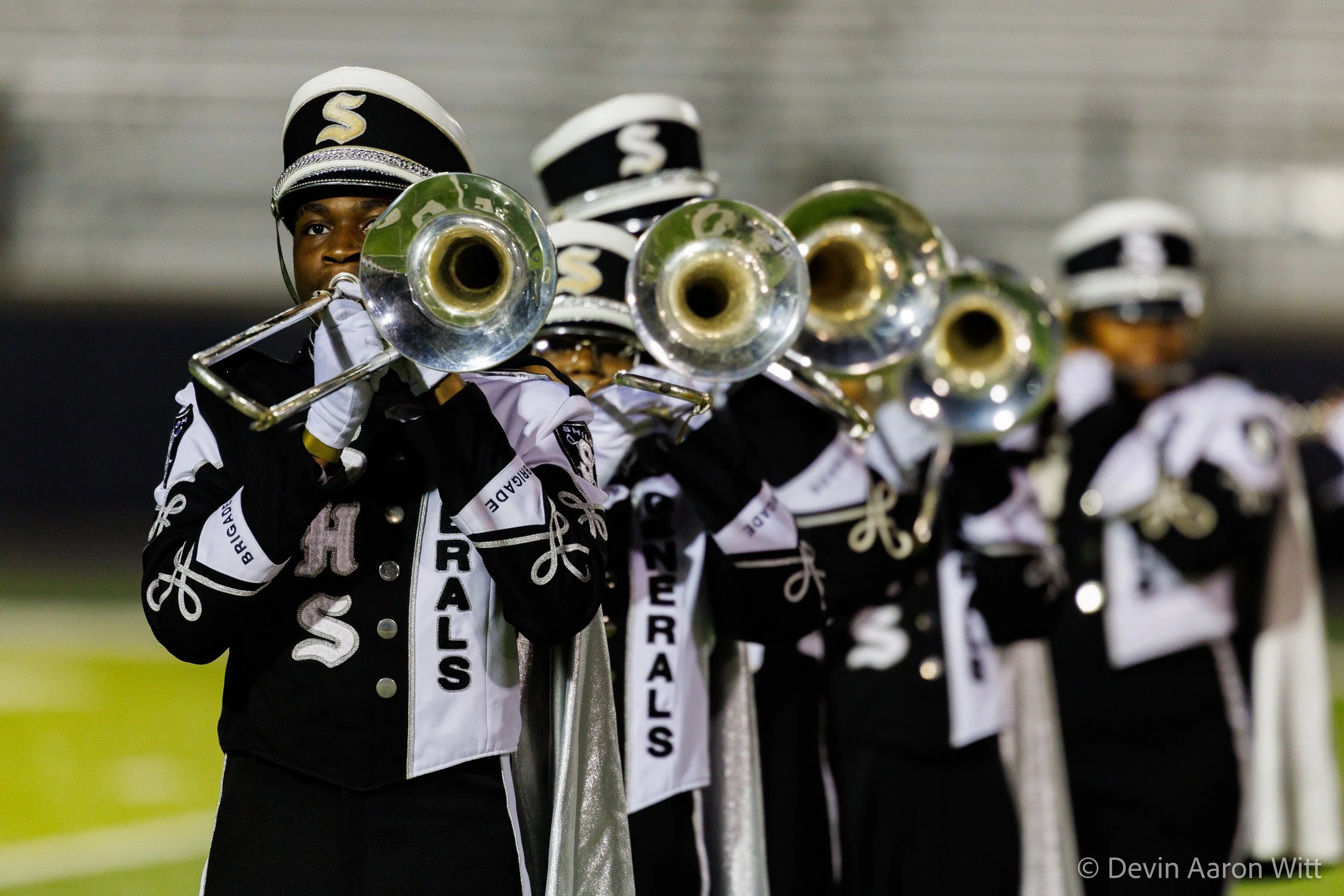High school marching band students in black and white uniforms playing brass instruments on the field during a nighttime performance.
