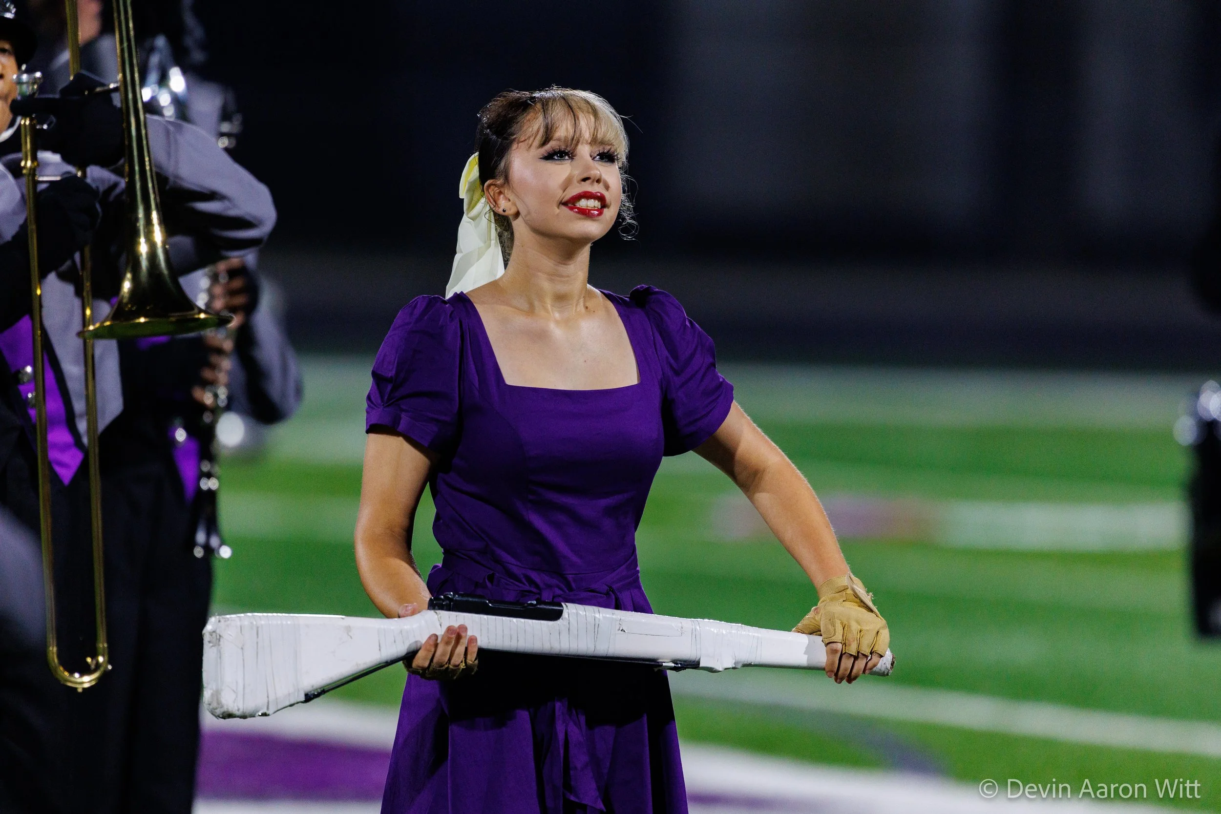 A woman in purple dress holding a baseball bat, standing on a football field during a night game, with band members and instruments around her.