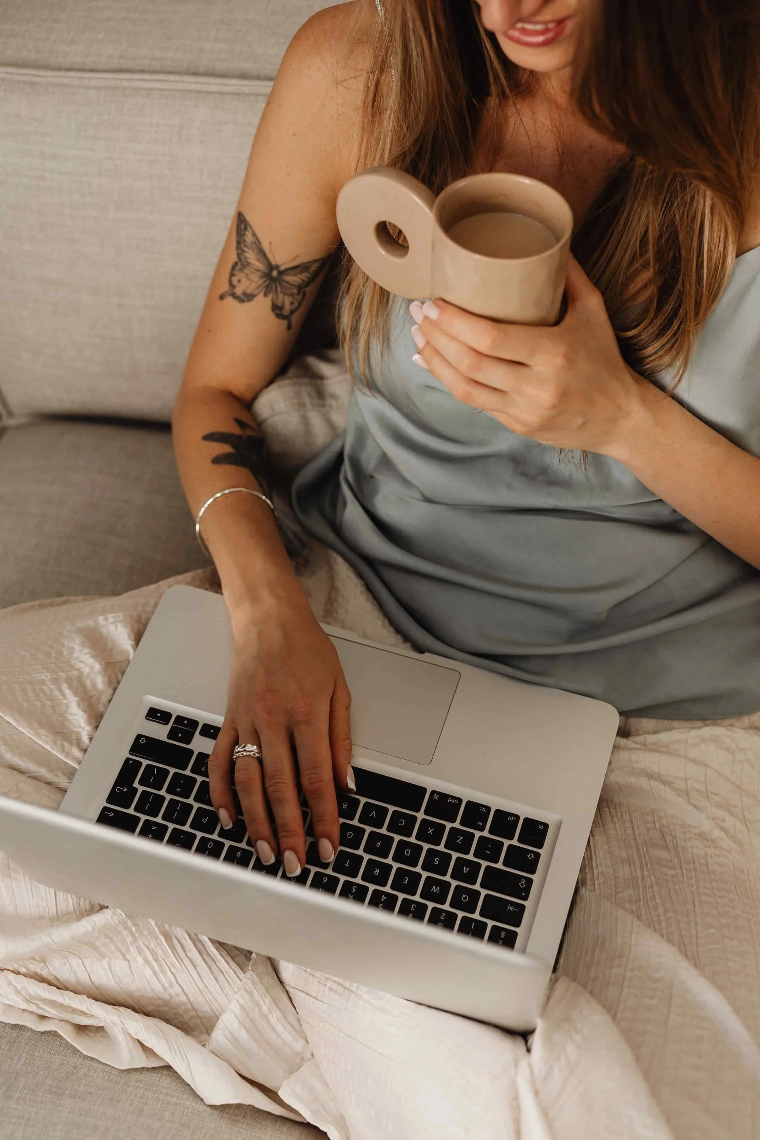 A woman with long brown hair and tattoos on her arms, wearing a gray silk camisole, sitting on a couch with a laptop on her lap. She is holding a beige mug with both hands, and her nails are painted white. She is smiling slightly and looking down at her laptop.