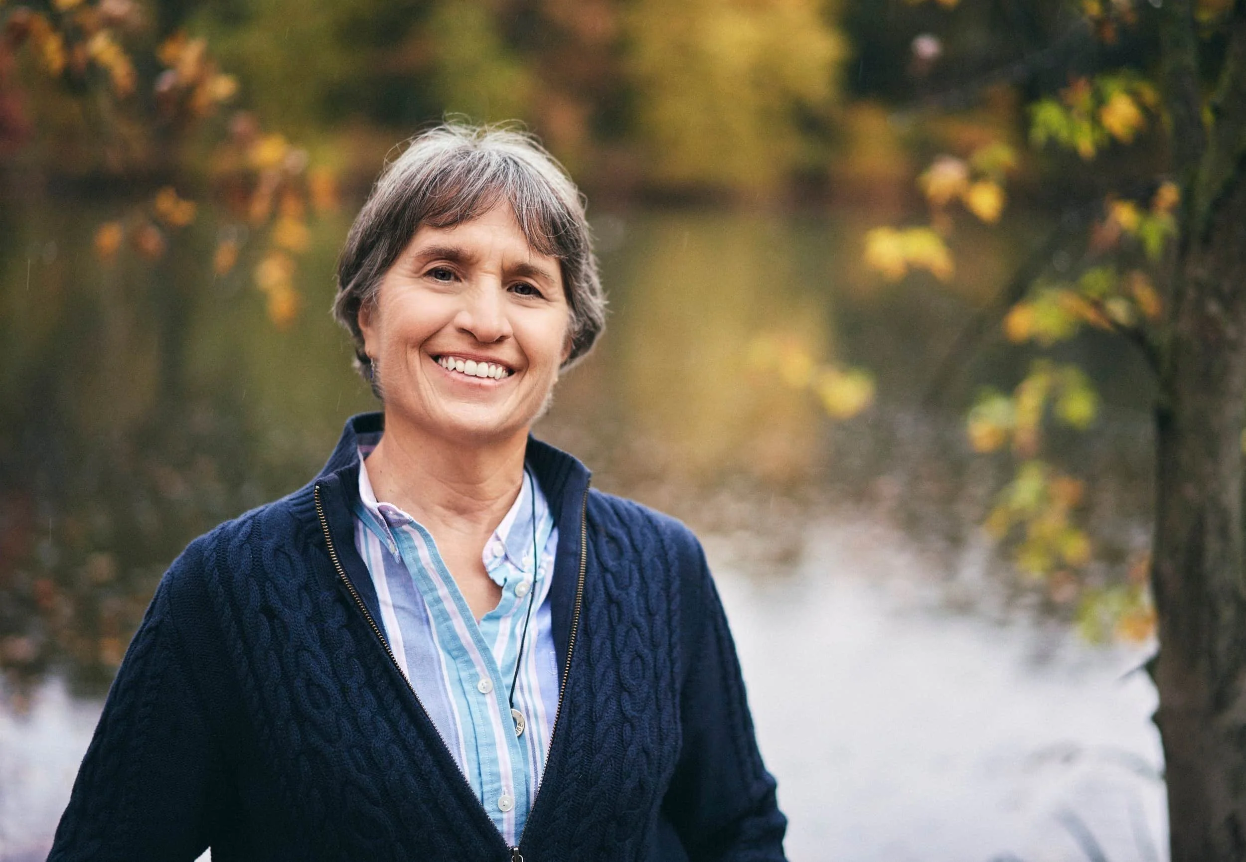 A middle-aged woman with short gray hair smiling outdoors near a river with trees in autumn foliage.
