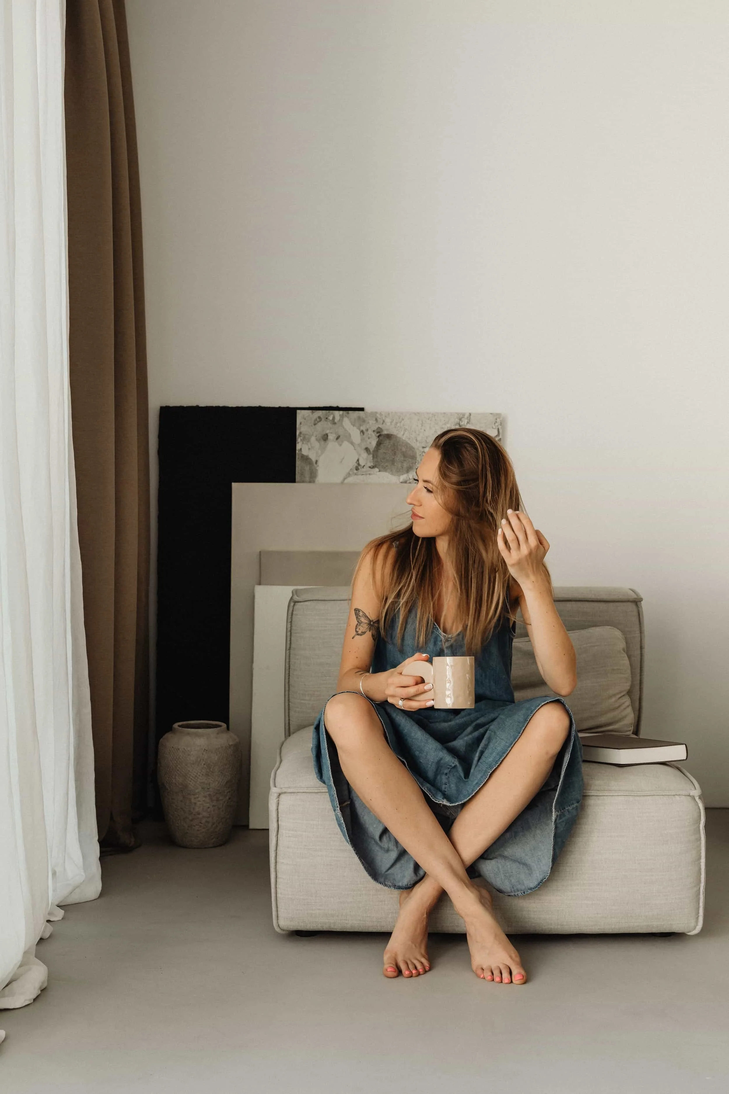 A woman with long, wavy hair, tattoos on her upper arm, sitting cross-legged on an armchair, holding a beige mug, and looking to her left in a room with neutral decor and a large abstract headboard.