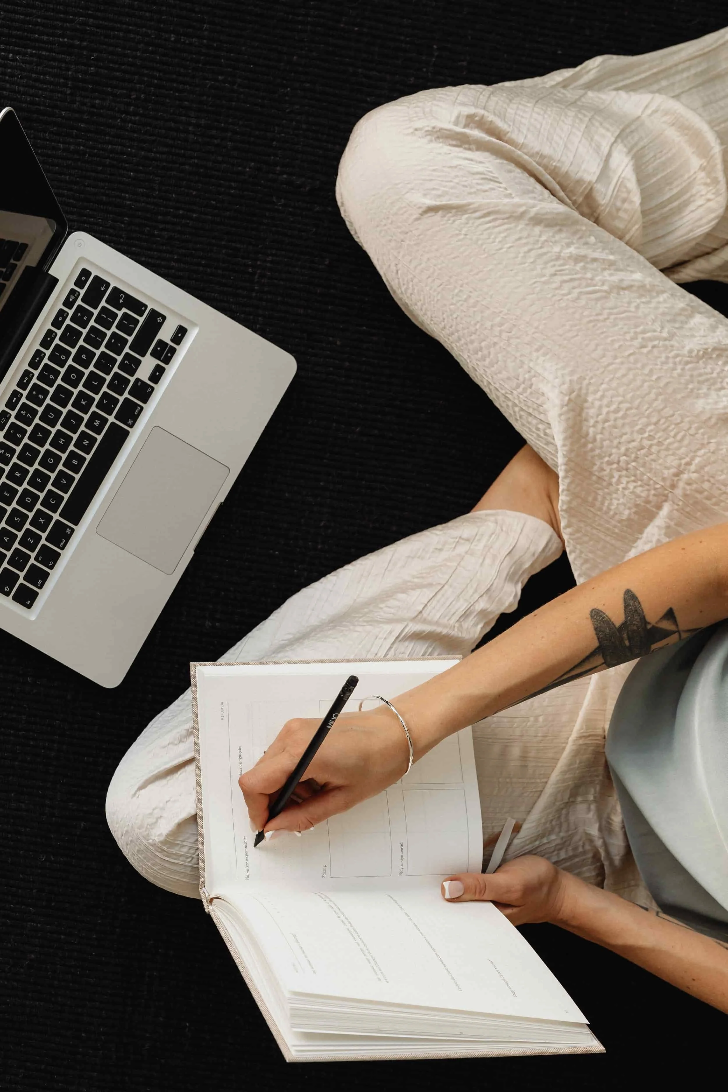 A person in a white dress writing in a paper notebook, with a laptop nearby, sitting on a black textured surface.