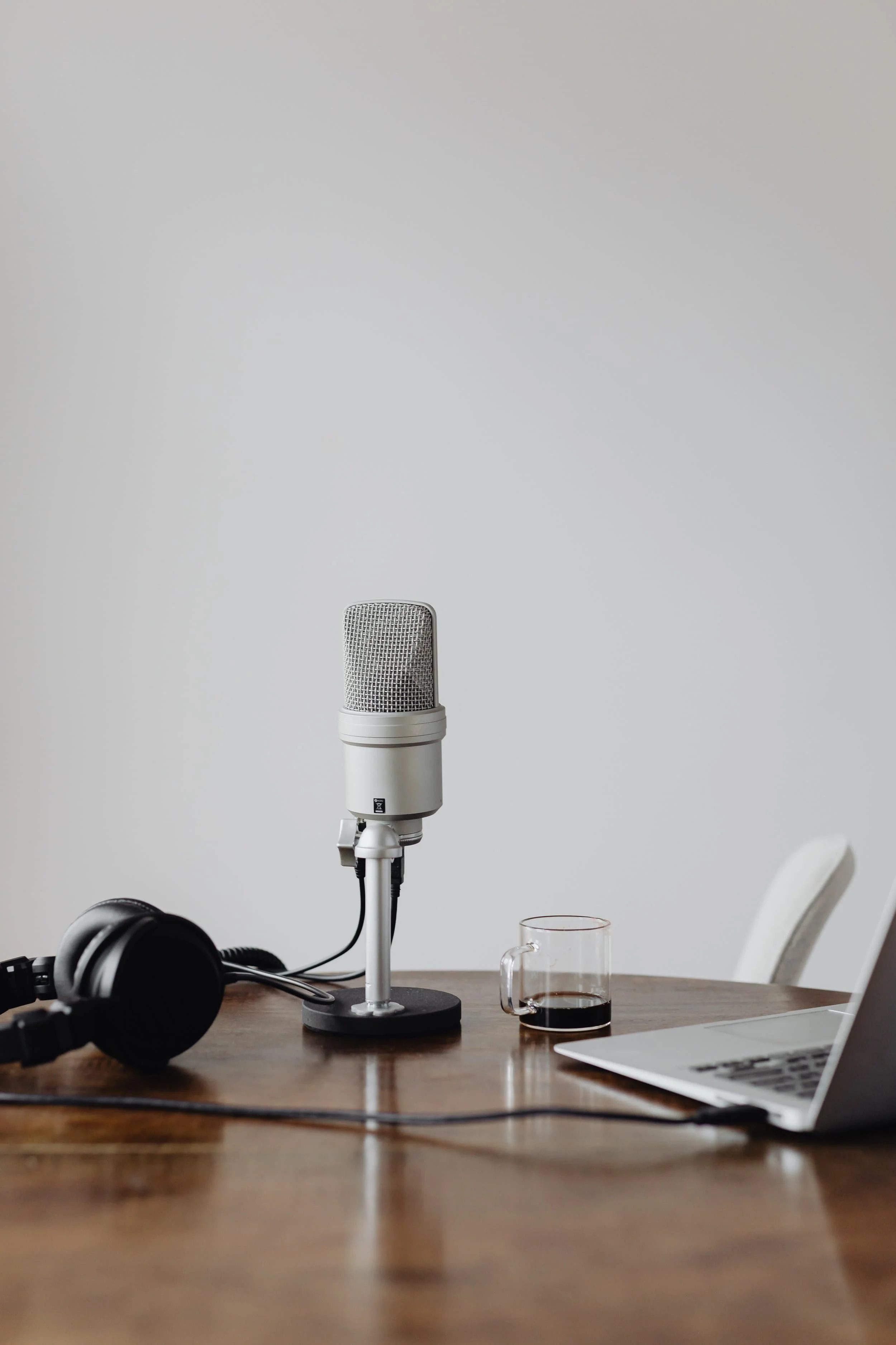 A white microphone on a stand, a glass mug filled with dark coffee, and a pair of black headphones on a wooden table, with a computer keyboard in the bottom right corner and a gray chair in the background.