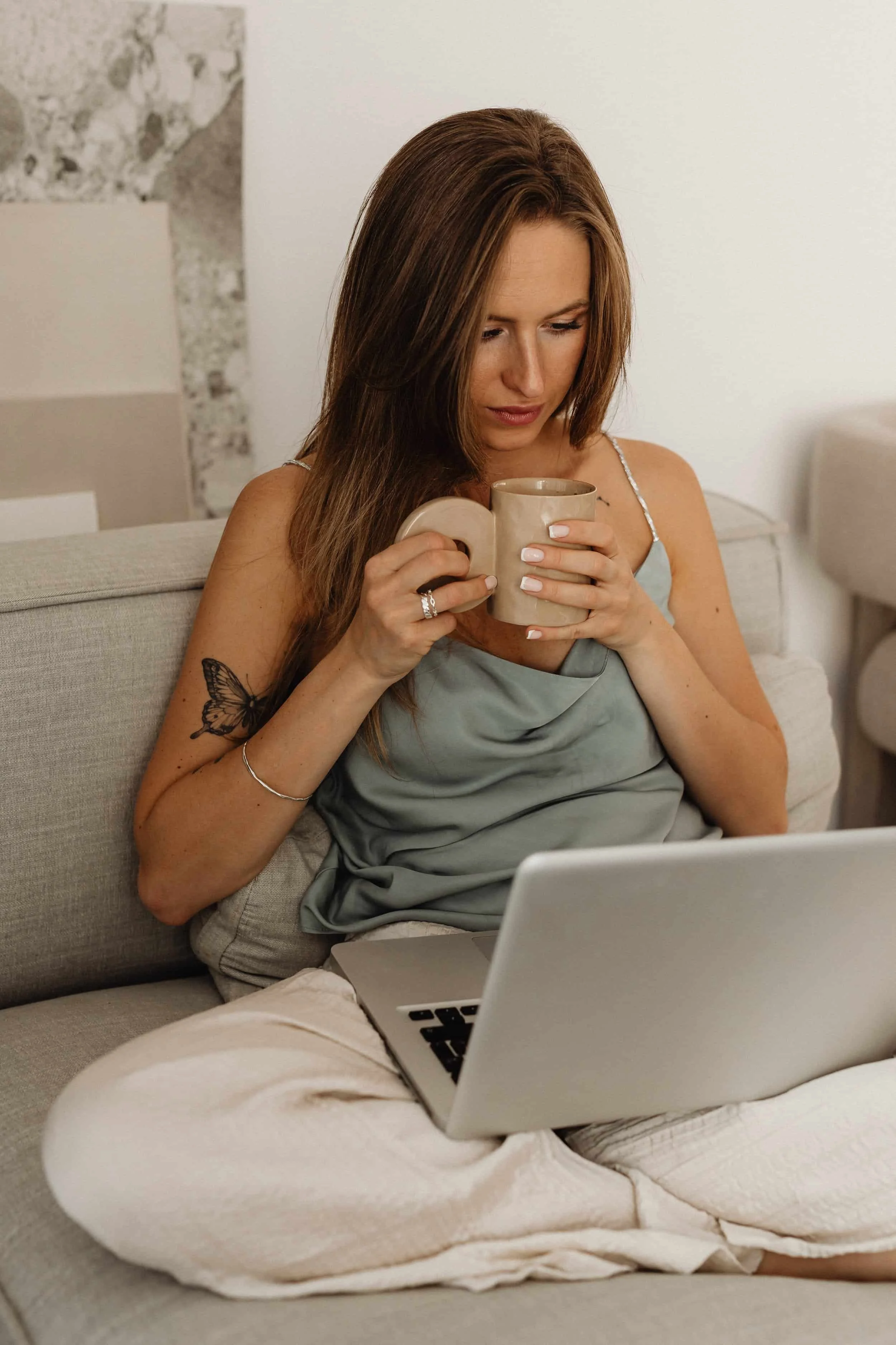 Woman with brown hair, tattoos, and jewelry, holding a coffee mug while sitting on a beige couch with a laptop on her lap.