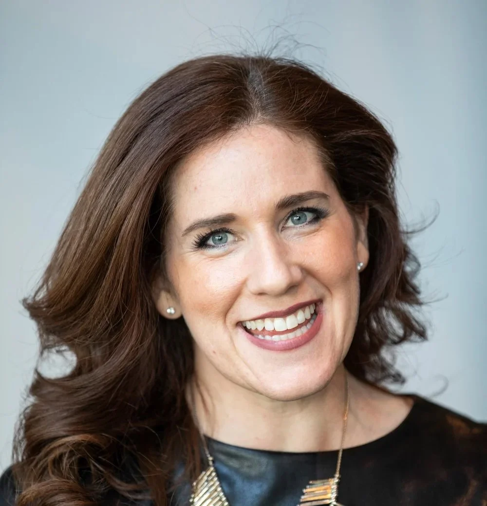 A woman with shoulder-length brown hair and blue eyes smiling, wearing pearl earrings and a black top with gold jewelry.