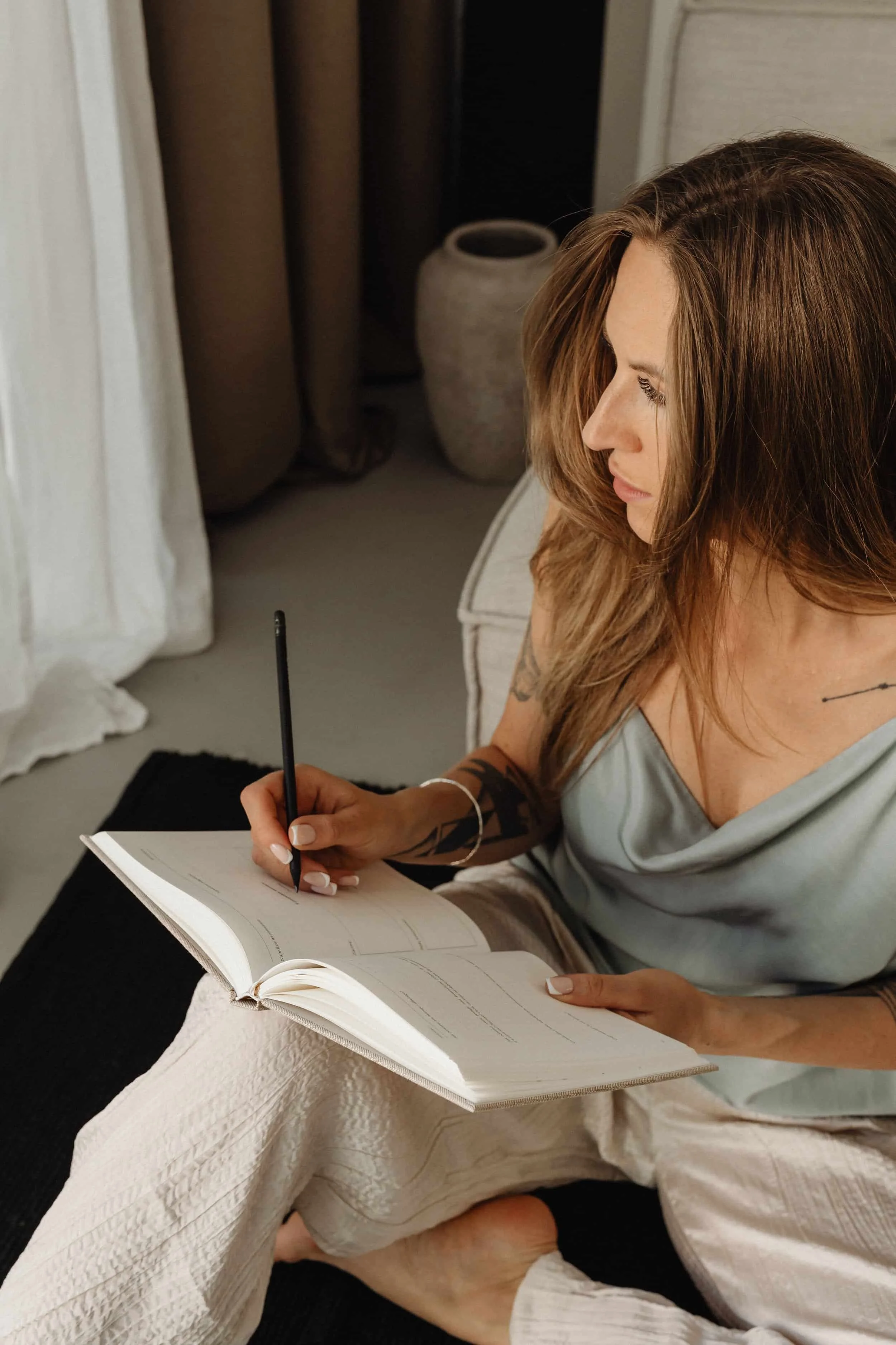 Woman with brown hair and tattoos sitting on a bed, writing in a notebook, with a large vase and curtains in the background.