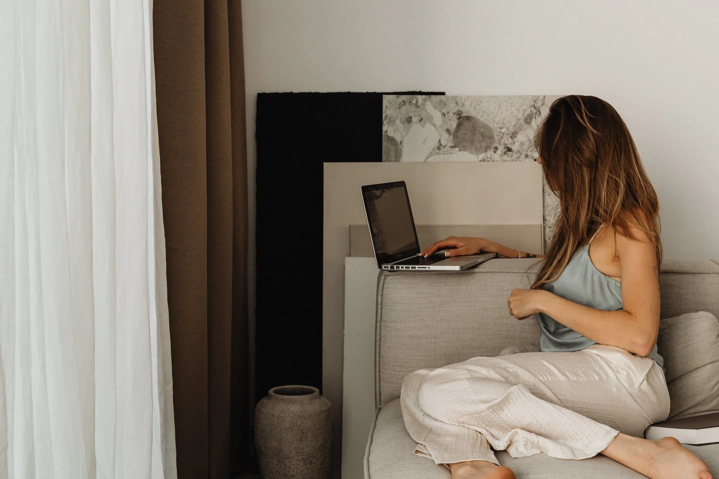 A woman with long hair sitting on a beige sofa, using a laptop on her lap in a room with white walls and brown curtains.