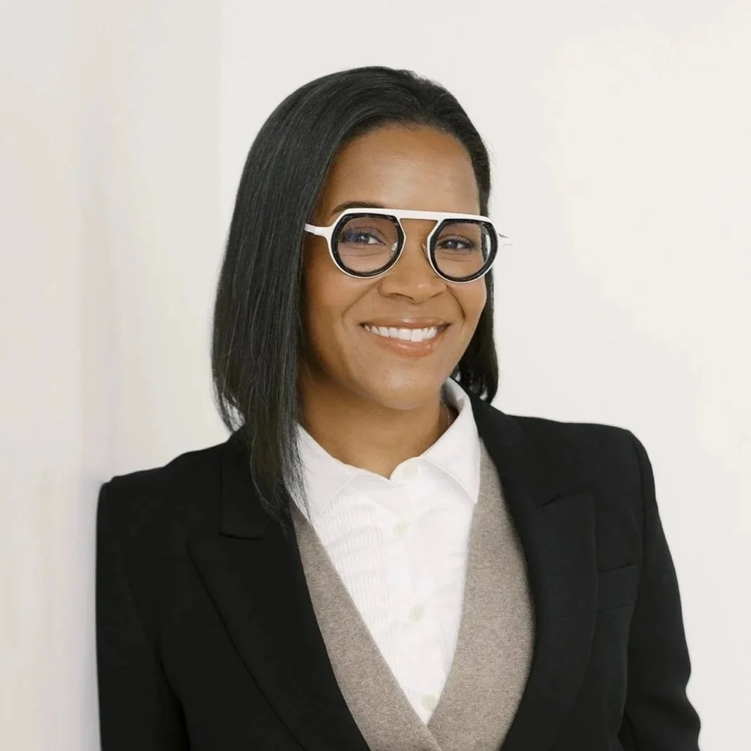 A professional woman with shoulder-length black hair, wearing glasses, a white collared shirt, a beige vest, and a black blazer, smiling, standing against a plain white background.