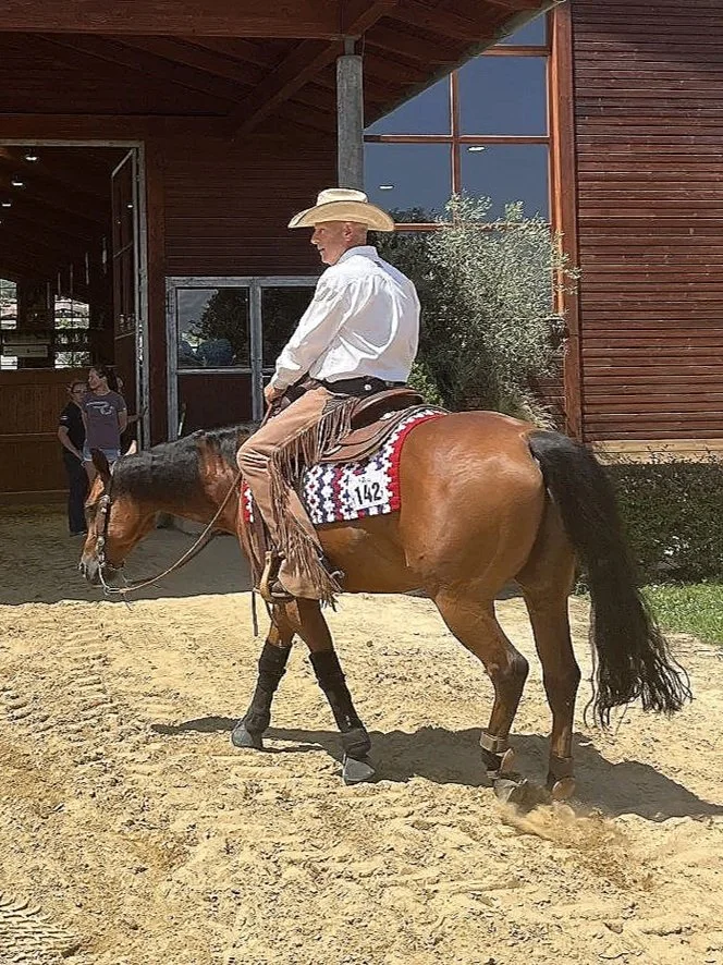 A cowboy riding a brown horse with a black mane and tail, wearing a white cowboy hat, white shirt, and fringed chaps, standing on sandy ground outside a wooden building with large windows.