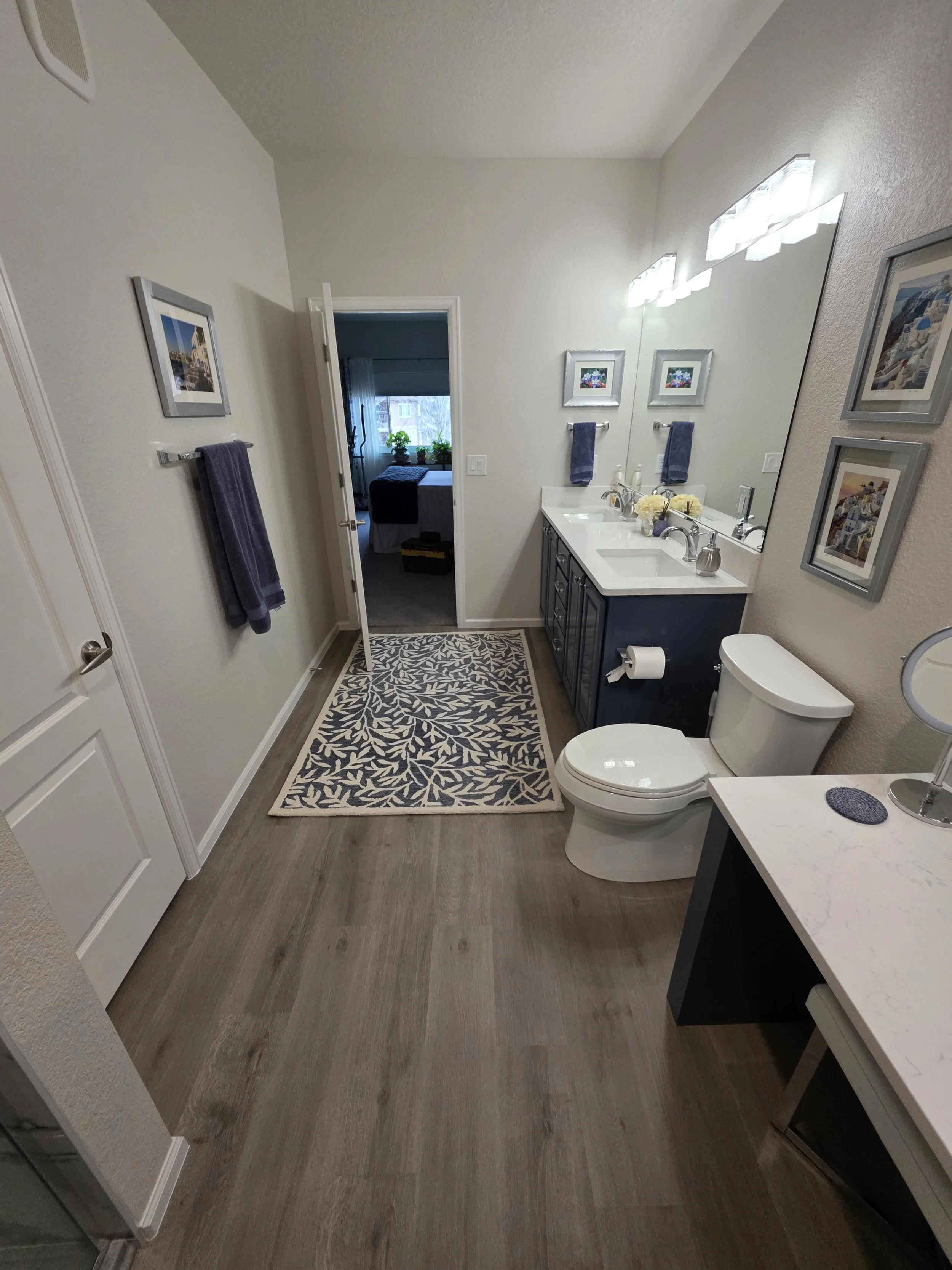 A clean and modern bathroom with a double vanity sink, a mirror, a toilet, and wall art. There are towels hanging on the wall and a patterned rug on the wooden floor. An open door leads to a bedroom with a window and plants.