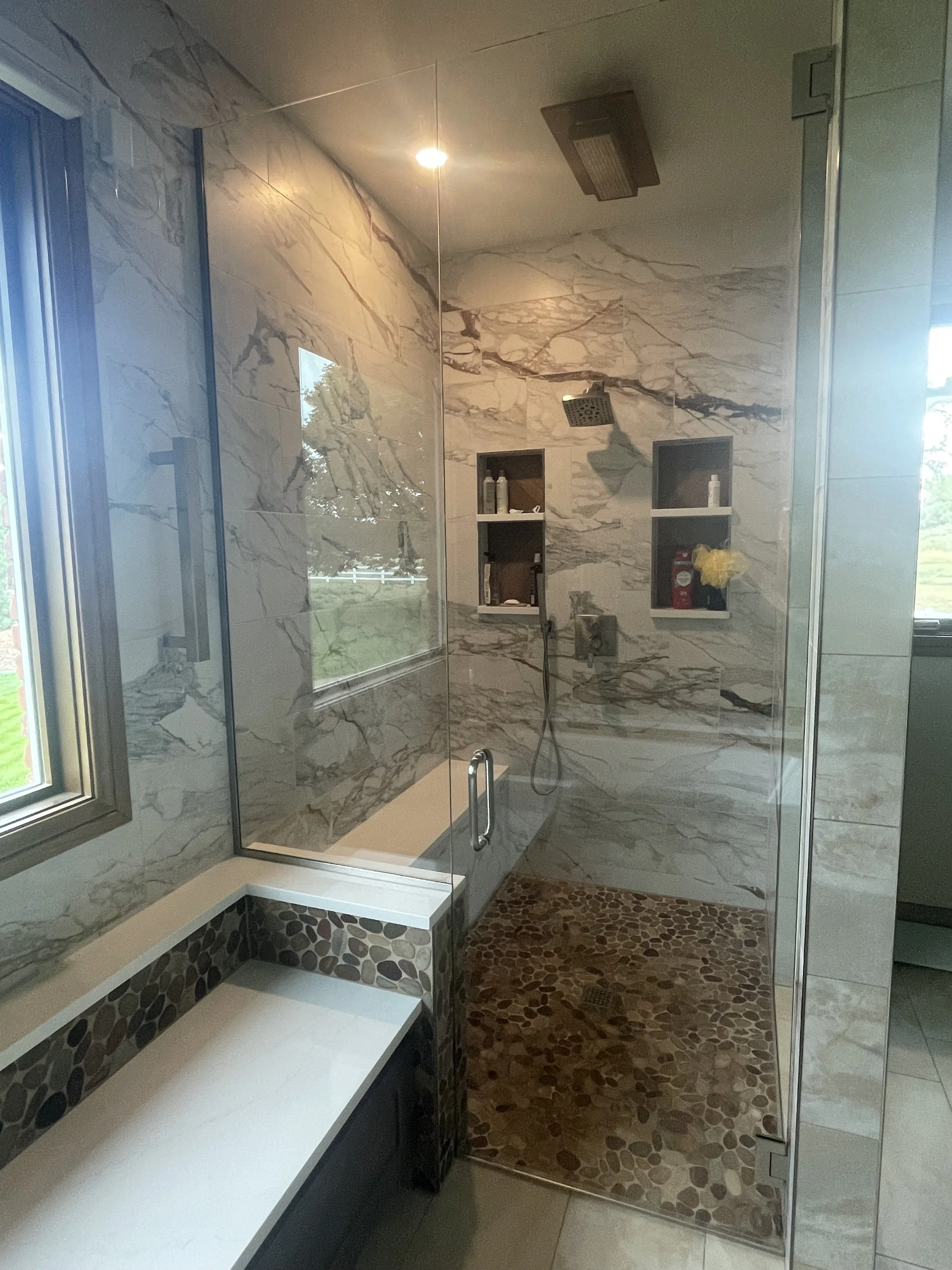 Bathroom shower area with marble walls, a bench with pebble design, a built-in shelf with toiletries, and a window with a view of trees and a fence.