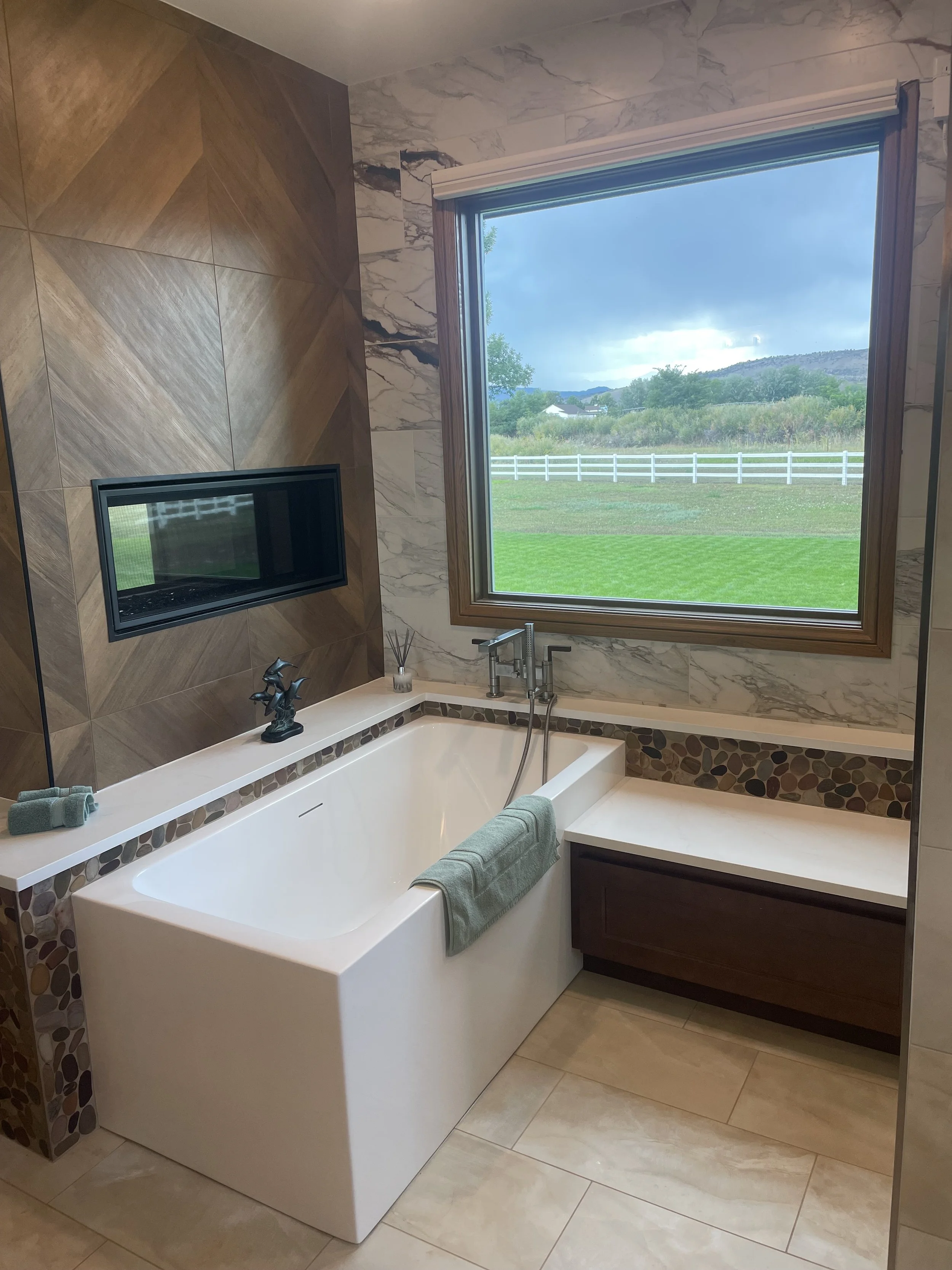 A modern bathroom with a large soaking tub and a view of the countryside through a big window. The bathroom features marble and wood accents, with a small sculpture on the tub's ledge, and a green towel hanging on the tub.