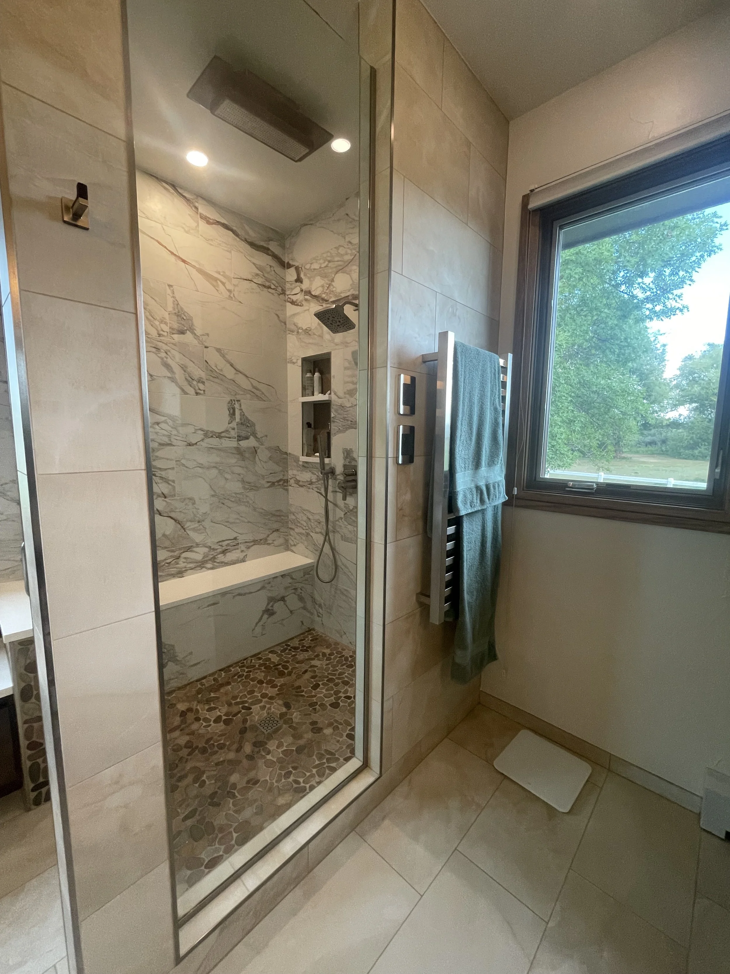 Bathroom shower with marble tiles, a built-in niche for toiletries, a rainfall showerhead, and a glass door. There is a window showing green trees outside, a towel hanging on a towel rack, and a digital scale on the floor.