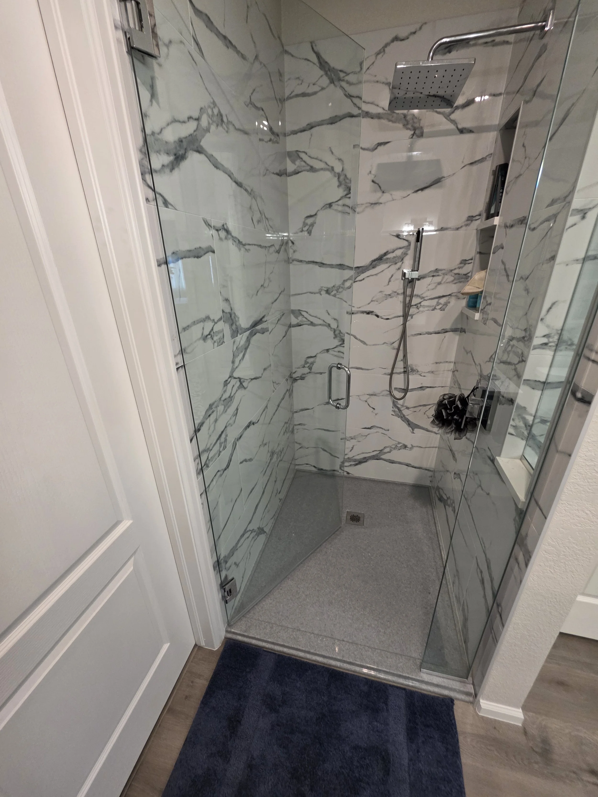 A modern walk-in shower with clear glass doors, gray marble walls, a rain shower head, a handheld shower, and built-in shelves, with a dark blue rug on the bathroom floor.
