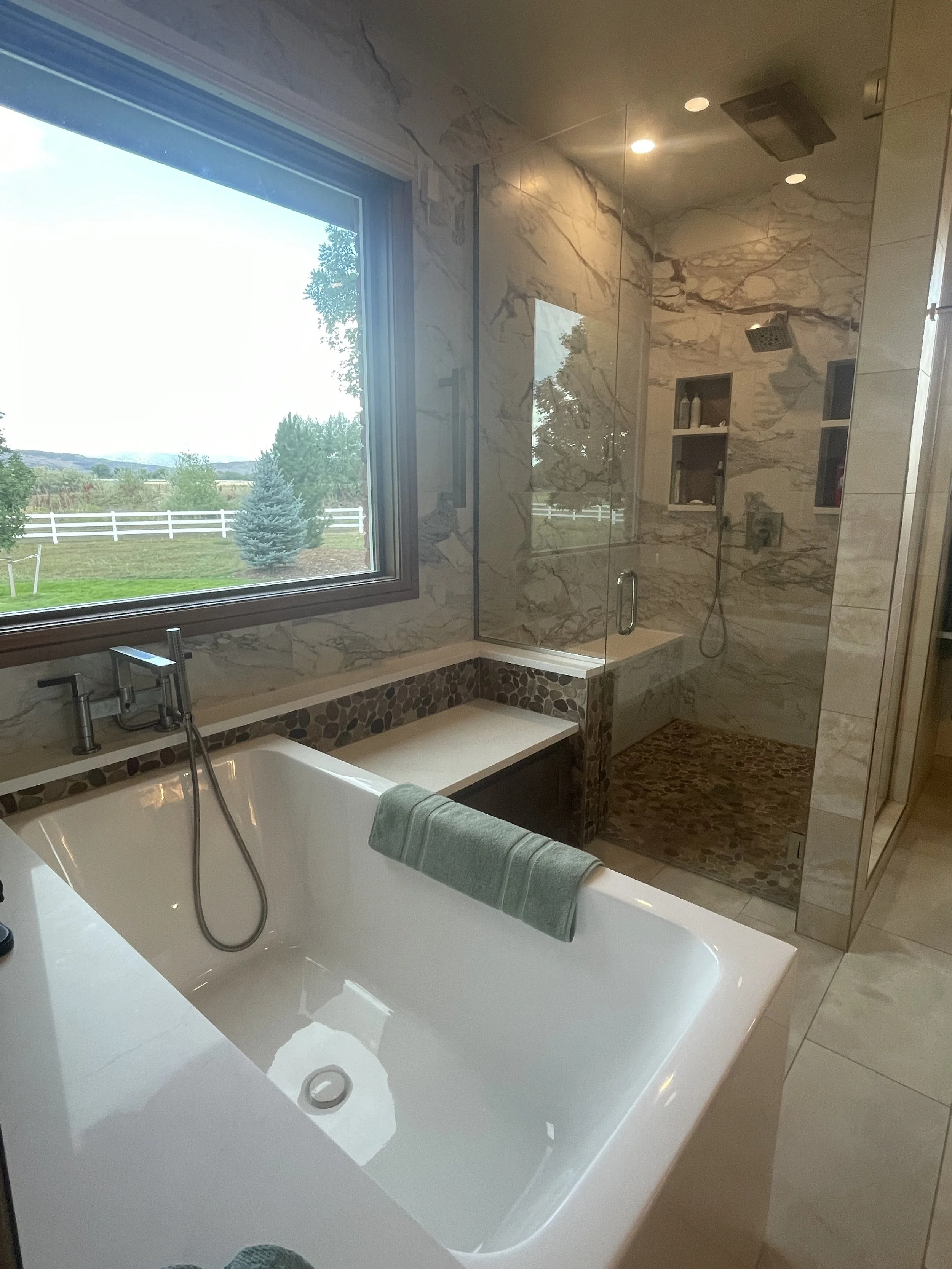 A bathroom with a large window showing an outdoor view of trees and a white fence. The bathroom features a marble shower with glass doors, built-in shelves, and a rainfall showerhead. There is a bathtub with a green towel draped over it.