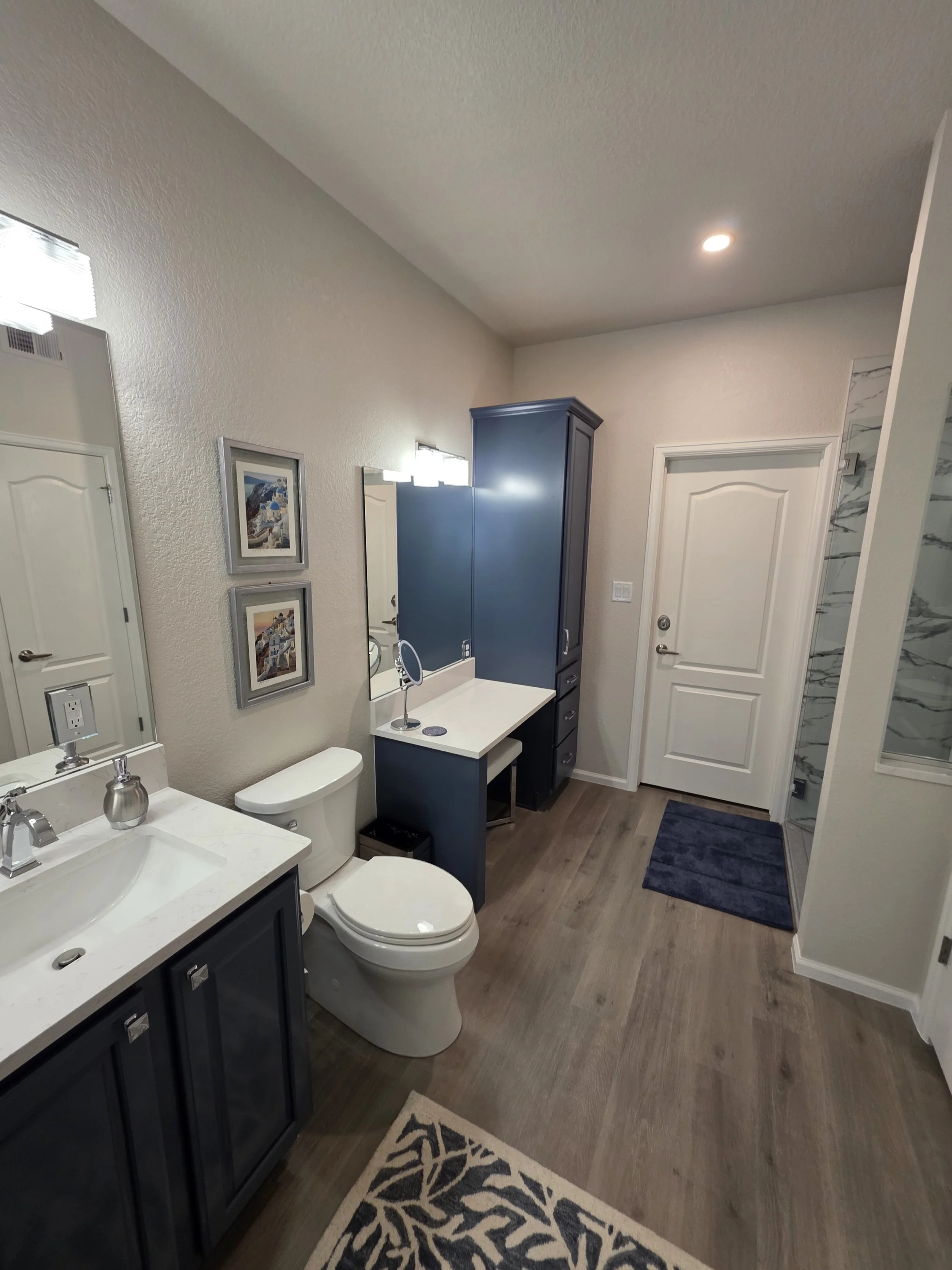 Bathroom with dark vanity, white countertop, mirror, toilet, blue cabinet, small desk with mirror, framed pictures, dark blue rug, wooden floor, shower with marble tiles.