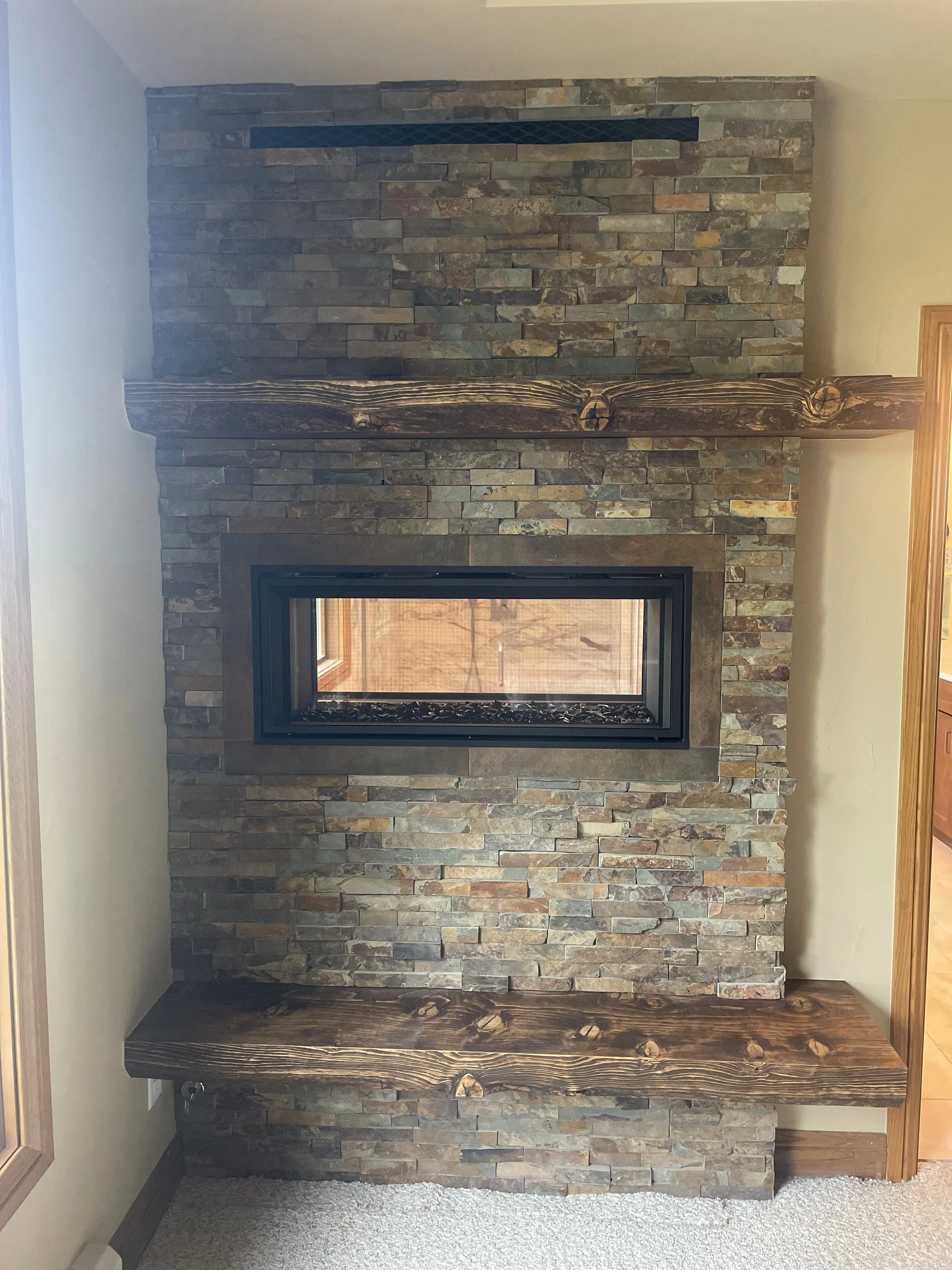 A decorative fireplace with a black metal insert, surrounded by a stone veneer wall. There is a wooden mantelpiece on top, and the floor is carpeted.